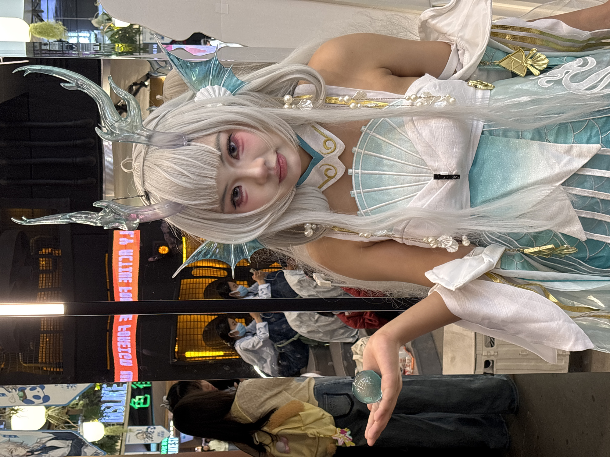 A young woman in a cosplay outfit is posing, holding a small blue object. The cosplay includes a white wig with horn-like accessories, a blue and white costume, and decorative elements. The scene appears to be indoors, in a public space, possibly a shopping mall or convention center. Reflections in the background suggest other people are present, and store signs are visible. The city is Liangjiang, China.