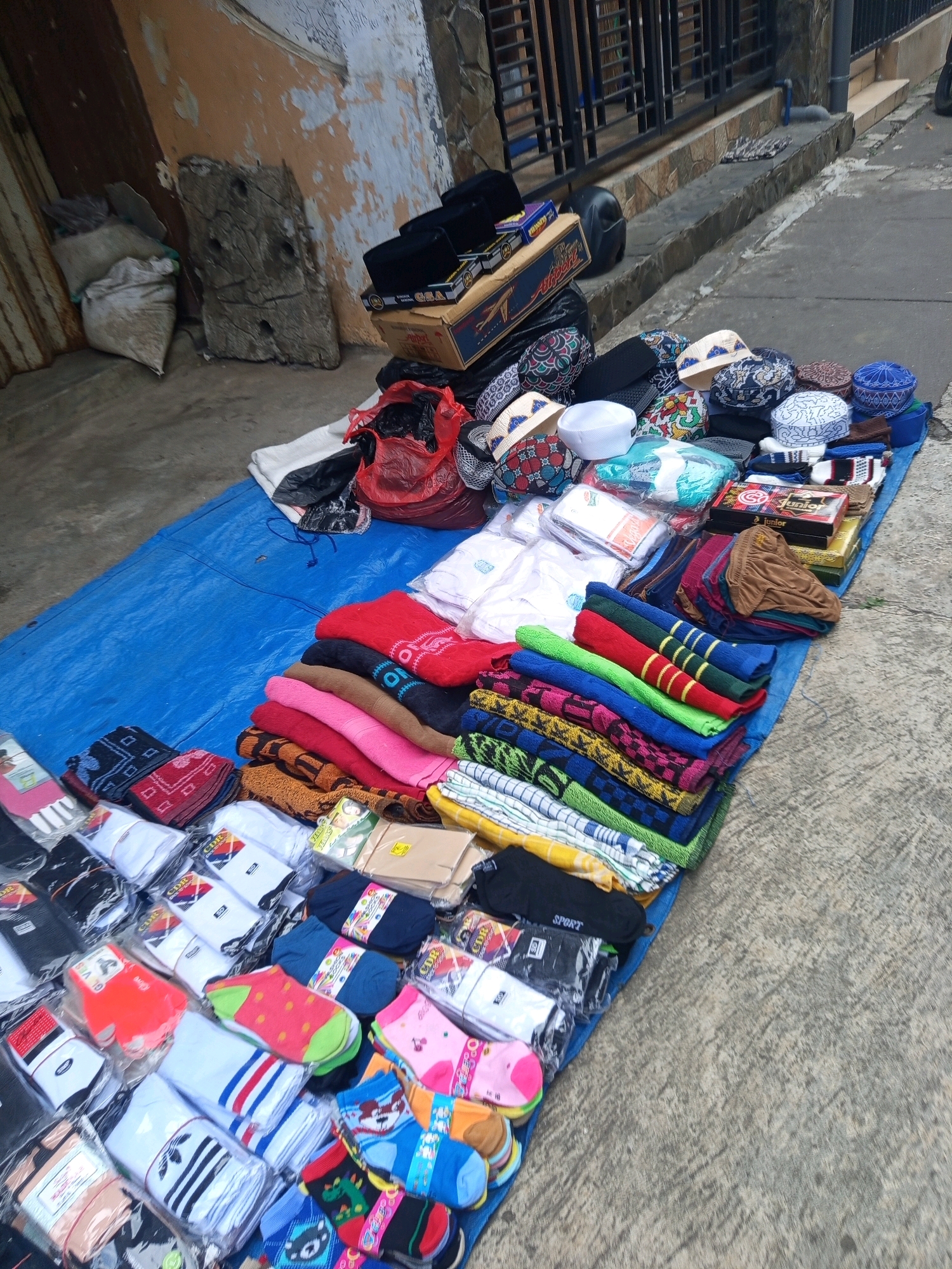 An outdoor street stall displays various religious and clothing items, including prayer caps, socks, and folded garments, spread on a blue tarp on a concrete sidewalk. The stall is located in front of a building with a stone facade and a metal fence. The items for sale are neatly arranged in stacks and piles. Some items are packaged, while others are folded. In the background, a wooden object and bags are visible next to the building. The scene appears to be in an urban environment, possibly in the city of Malang, Indonesia, given the architectural style and the type of merchandise. There are no people visible in the frame.