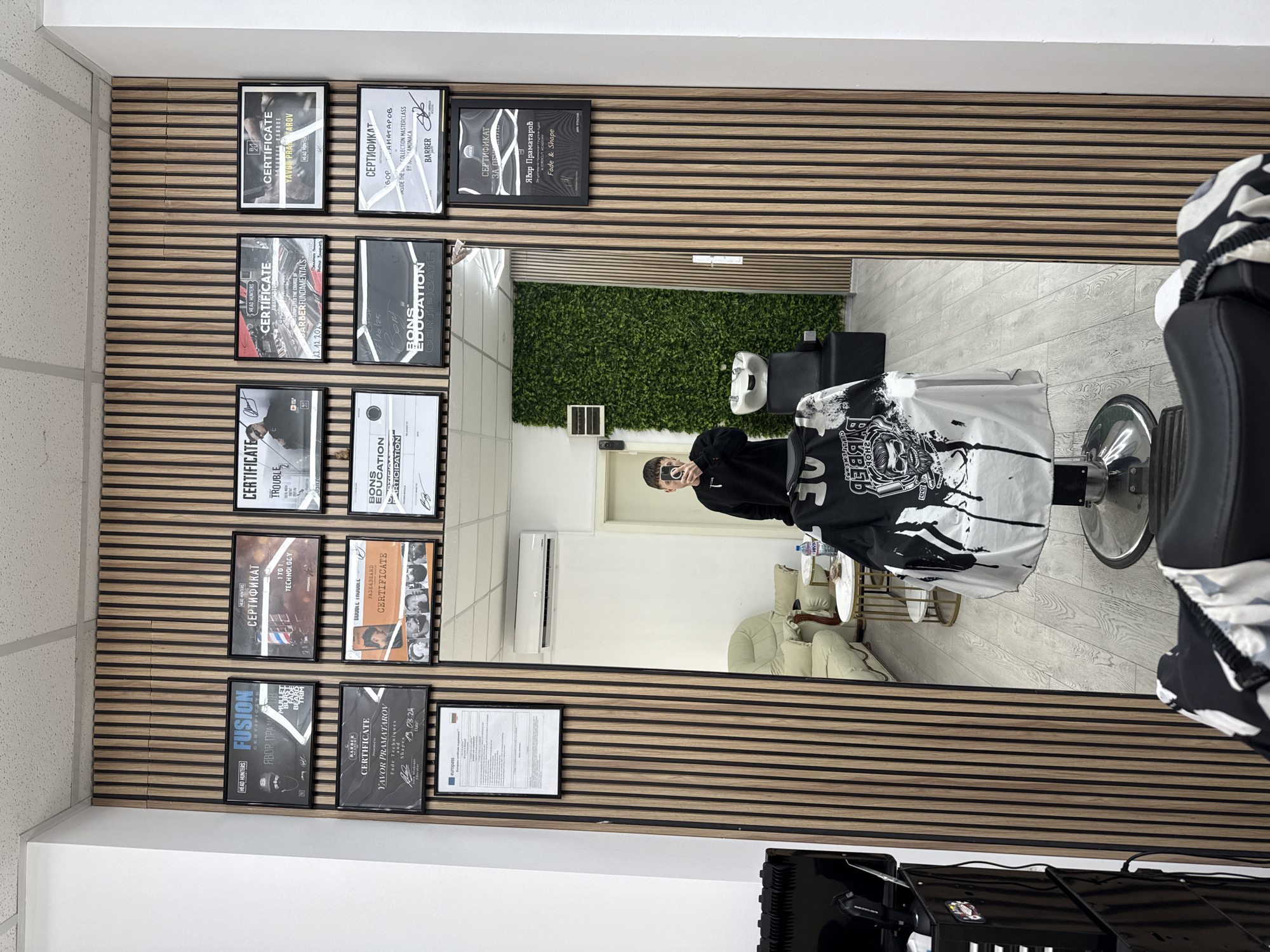 The image captures the interior of a modern barbershop located in Sofia, Bulgaria, offering a glimpse into a professional and well-equipped establishment. A large mirror, occupying a significant portion of the right side, reflects a person taking a selfie with a smartphone, likely the barber or owner, dressed in a dark long-sleeved top. The reflection also shows a black barber chair with a distinctive black and white patterned cape draped over it.
The main wall visible on the left is a stylish wooden slat design, serving as a display for numerous framed certificates and diplomas. These documents showcase the barber's extensive training and expertise, with visible names like "Yavor Pramatarov" (Явор Праматаров) and organizations such as "Head Hunters" and "Bons Education." Specific skills and courses mentioned on the certificates include "Barber Fundamentals," "Fade & Shape," "1 to 1 Technology," "Beard Trim," "Mullet Fade & Beard," and "Inside the Collection Masterclass by Jamonaca." Dates like "19.03.24," "22.12.2023," and "7/10/25" are also present. One framed document contains the word "EUROPAS."
Further reflected in the mirror are other elements of the barbershop's interior: a vibrant green artificial plant wall, a white wall with an air conditioning unit mounted high, and a cozy waiting area furnished with light-colored armchairs and a small golden-framed table. The floor throughout the shop appears to be covered in light wood-effect tiles.
