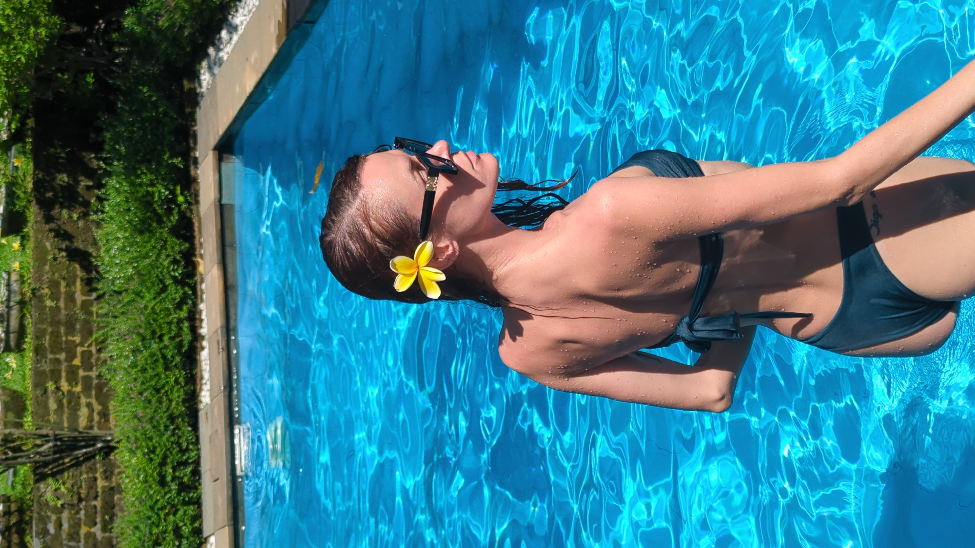An overhead, slightly angled shot captures a woman relaxing in a vibrant blue swimming pool, set in a sunny, tropical environment, likely North Kuta, Indonesia. The woman, viewed from her upper body, is floating on her back, wearing dark sunglasses and a dark-colored two-piece bikini. Her brown hair is wet, and a bright yellow plumeria flower is tucked behind her right ear. Her skin glistens with water droplets, indicating recent immersion.
The swimming pool features clear, brilliant blue water with distinct light refractions and ripples on its surface, reflecting bright sunshine. The pool's edge has a light-colored coping. To the left of the pool, lush green foliage, possibly a garden or dense bushes, and a textured stone or brick wall are visible, reinforcing the outdoor, tropical setting. The strong shadows and bright illumination suggest it is daytime, likely under a clear sky.
The woman's posture conveys relaxation and tranquility as she floats effortlessly. A small, indistinct tattoo, possibly cursive text like "Oiga," is visible on her right hip/lower back area. The scene depicts a peaceful moment of leisure in a picturesque, sun-drenched location.