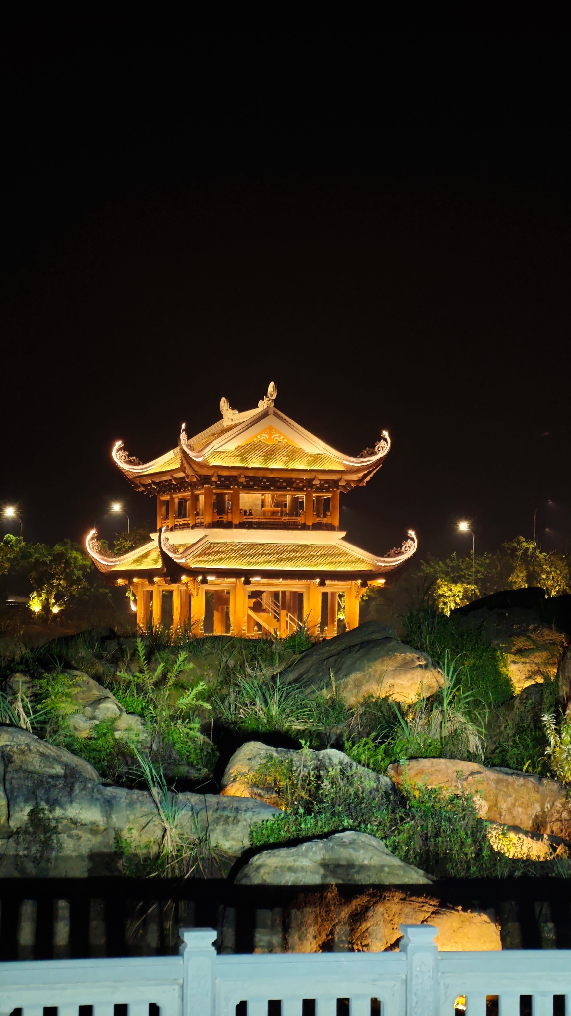 This is a nighttime, vertical, medium shot of a brightly lit, multi-tiered traditional Vietnamese pavilion situated on a rocky, overgrown embankment. The pavilion, with its ornate, upturned rooflines illuminated by warm, yellow lights, stands as the central subject against a dark, night sky. The embankment is covered with various shades of green foliage and large, textured rocks, some of which are also subtly lit, highlighting their forms. In the foreground, a white, ornate railing is partially visible, suggesting a viewing area. Beyond the pavilion, faint lights from street lamps and trees are discernible, hinting at a surrounding park or urban landscape. There are no people or animals visible in the scene. The overall impression is one of serene, illuminated architectural beauty set within a natural, dark environment.