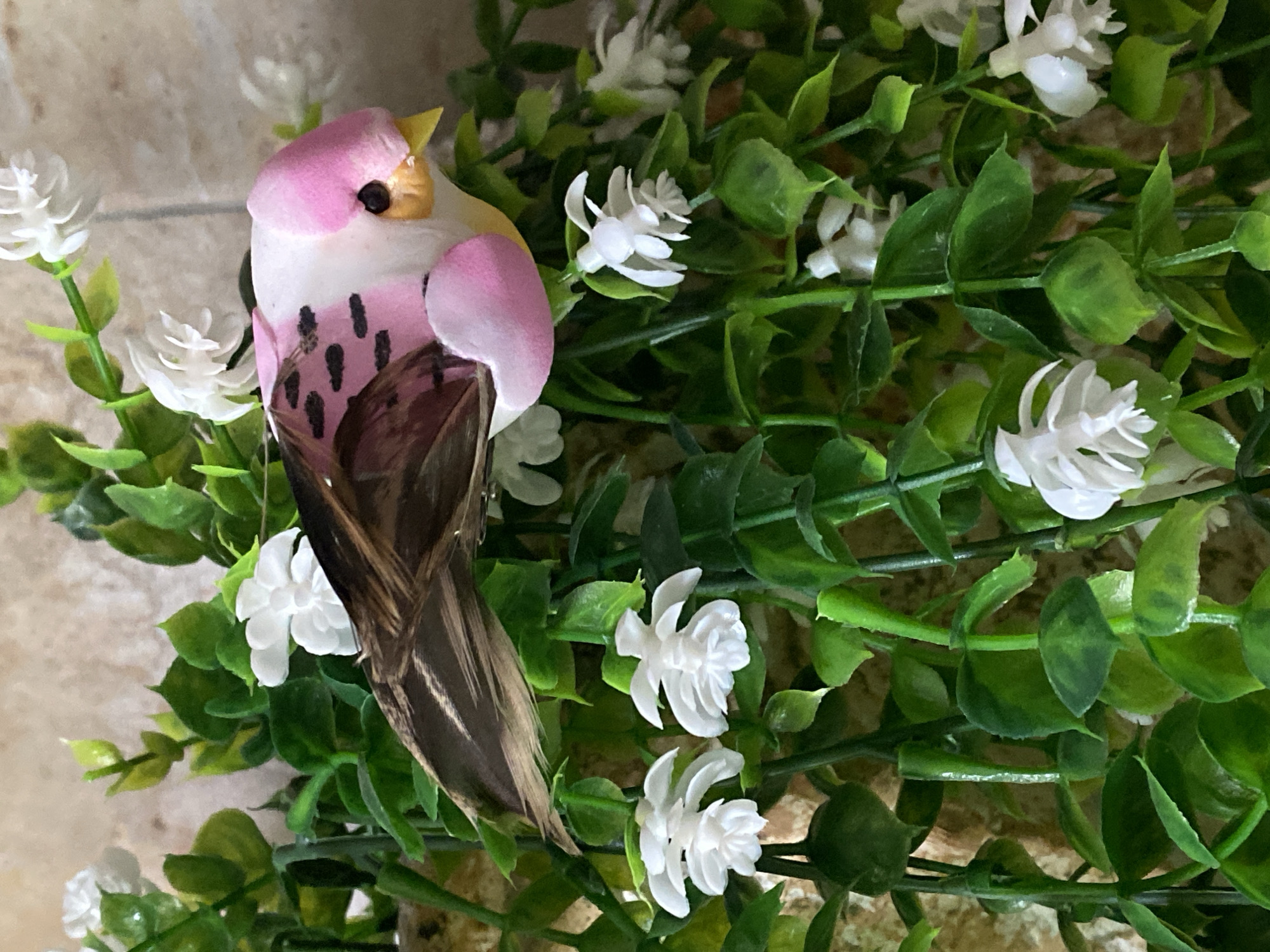 A close-up shot shows a decorative bird nestled among faux greenery and white flowers. The bird has a pink head, a white body with black markings, and brown and tan feathers for its tail. It has a yellow beak and a black eye. The greenery consists of small, round green leaves on thin, green stems. The white flowers are multi-petaled. The background is a neutral beige color.
The location in the city of Novokuznetsk, Russia cannot be ascertained from the image provided.