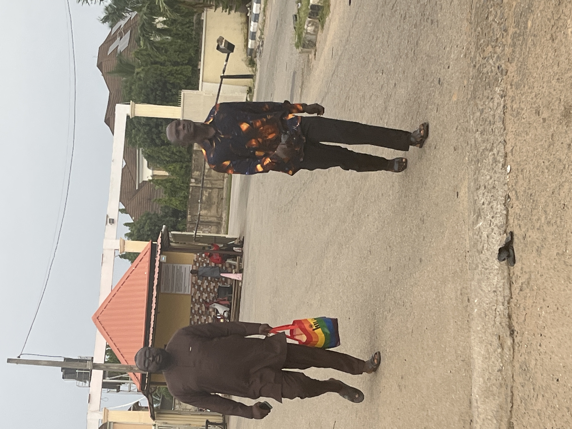Two men are walking on a paved road. The man on the left is wearing a brown agbada and sandals and carrying a rainbow-colored tote bag. The man on the right is wearing a black patterned shirt and black pants and sandals. In the background, there is a building with an orange roof, a gate, trees, and houses with solar panels. The sky is overcast. The scene appears to be in a residential area of Abuja, Nigeria.