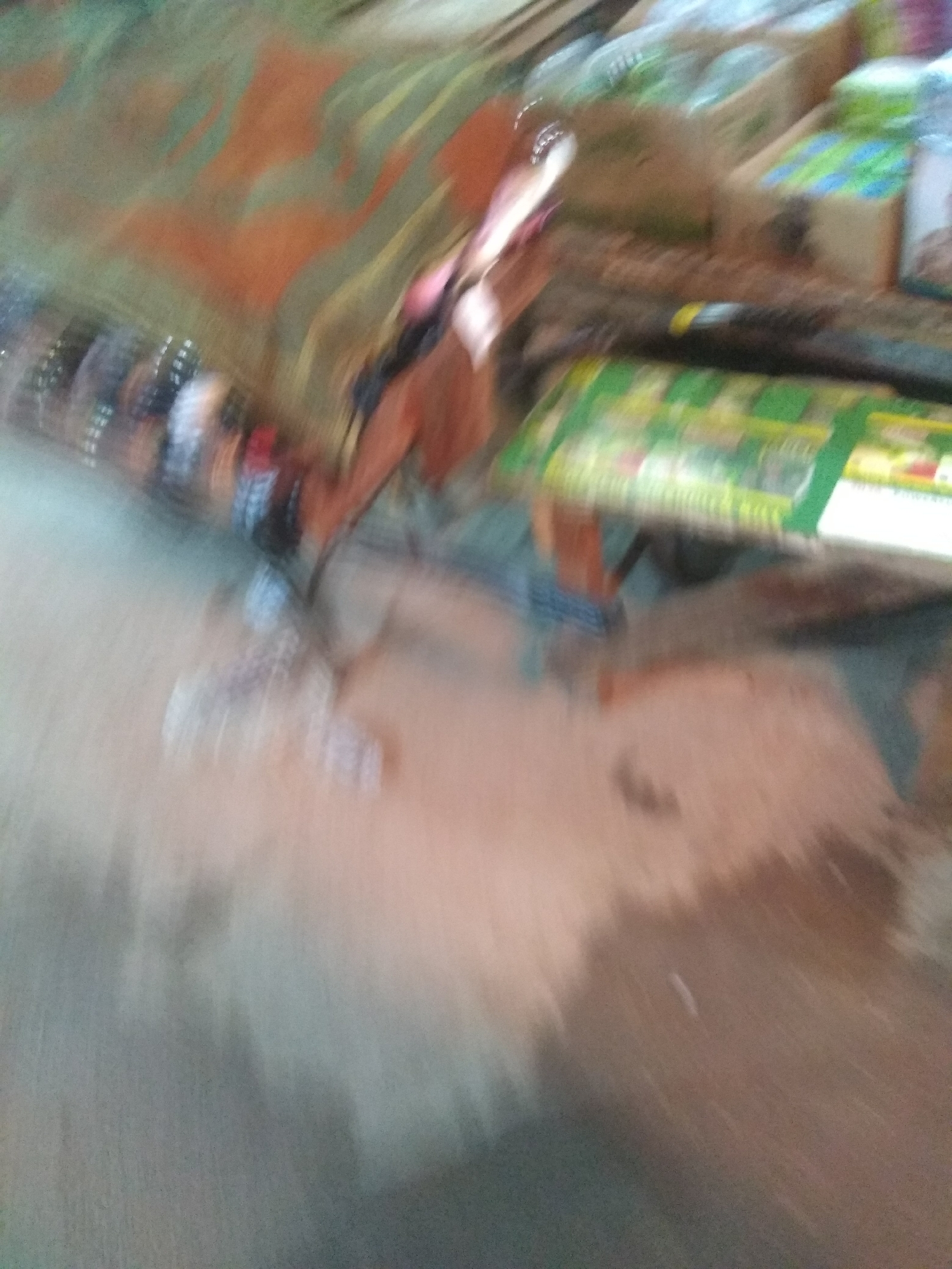 The image shows a blurry view of what appears to be a market or shop scene in Jalingo, Nigeria. The primary subjects are objects, possibly products for sale, and hints of human presence, though faces and clear figures are not discernible due to motion blur.
Objects visible include what seem to be bottles or containers on the left side, possibly in a row. In the center and to the right, there are boxes or packages, some with green elements, suggesting they might be food items, beverages, or household goods. The surface they are placed on is indistinct, but appears to be a table or stall.
The scene is characterized by significant motion blur, making it difficult to ascertain specific details about the activity or event taking place. However, the arrangement of goods and the presence of what might be packaging imply a commercial setting where items are being displayed or handled. The overall impression is one of a busy environment, potentially with movement contributing to the blur.
Notable details are scarce due to the image quality. The lighting suggests it could be daytime, but no specific time can be determined. There are no clear weather cues. The location context is provided as Jalingo, Nigeria. No discernible emotions are conveyed due to the lack of clear human subjects. There is no visible text that can be clearly read. The background is a mixture of colors and shapes, with swathes of orange, green, and beige, further contributing to the abstract nature of the image.