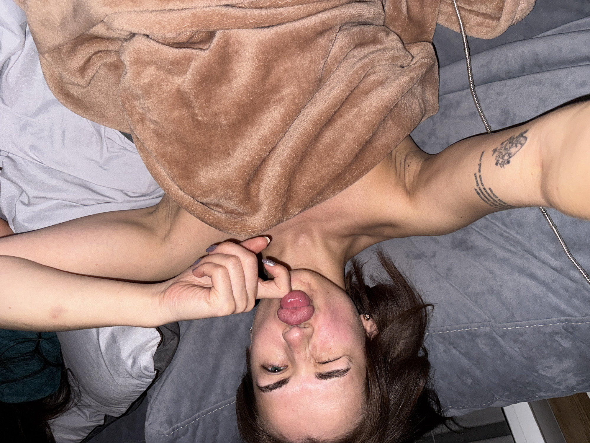 A young woman is lying on a grey bed with a brown blanket draped over part of her body. She is sticking her tongue out and touching it with her fingers. She has a tattoo on her right arm. The location is not discernible.