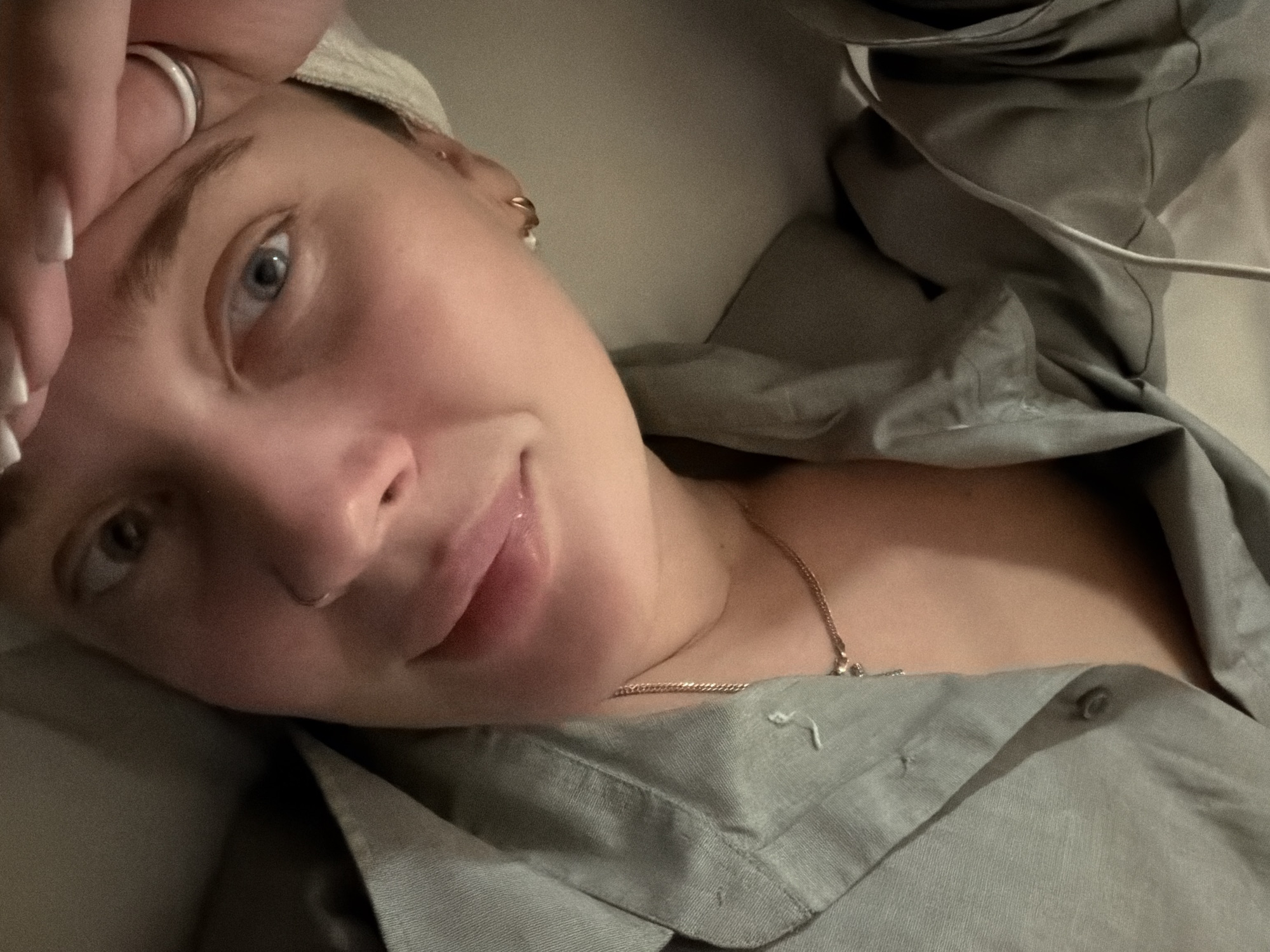 A close-up selfie depicts a woman with fair skin, light eyes, and short, possibly bleached hair. She's wearing a simple silver band on one of her fingers and a delicate gold necklace. Her top appears to be a grey linen or cotton shirt, unbuttoned at the top. A white cable is visible to the right of her shoulder, perhaps an earbud wire. The background is indistinct and light-colored. The subject is not located in Moscow, Russia.