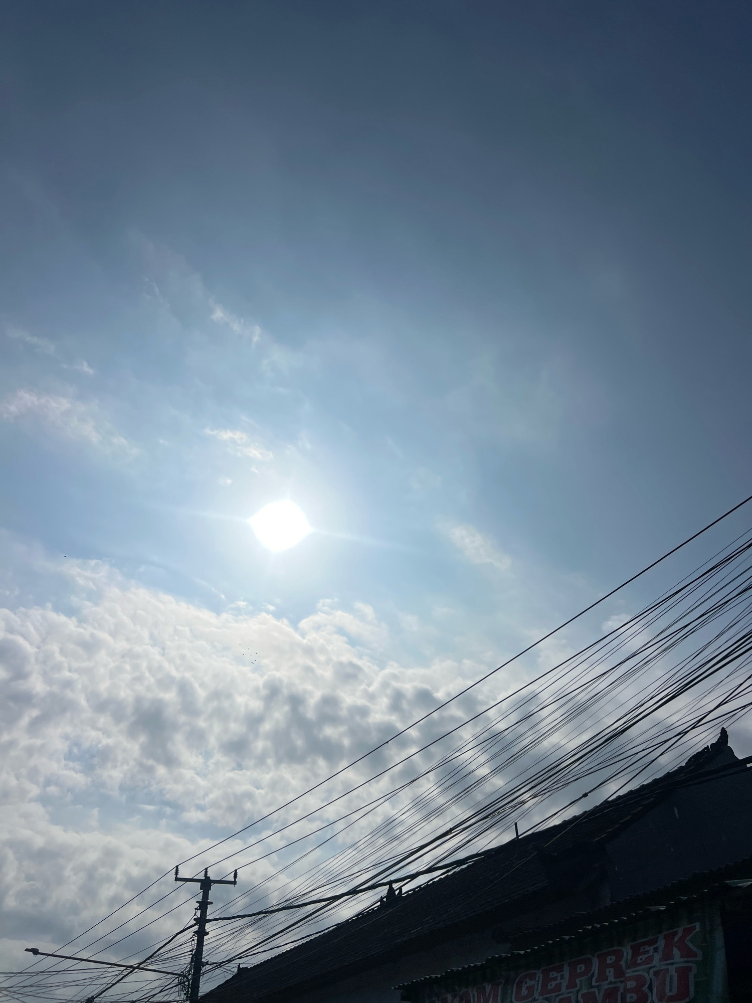 The image captures a bright, sunny day with a clear blue sky partially obscured by scattered white clouds. The sun is visible as a bright, blown-out orb near the center-left of the frame, emitting strong lens flares. In the foreground, the dark silhouette of a building's roofline is visible, topped with a dense network of black electrical wires. A utility pole with multiple crossbars stands prominently on the left side of the frame, also laden with wires. A sign with red lettering is partially visible at the bottom edge of the frame, with some of the letters appearing to spell out "AYAM GEPREK SAMBU". The overall impression is an urban scene, likely within a residential or commercial area of Denpasar, Indonesia, characterized by overhead power lines and the sun shining brightly.