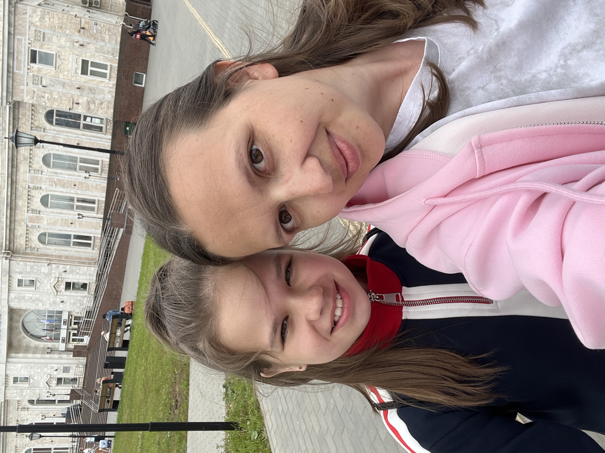 The image captures two females, an adult and a child, posing for a selfie in front of the Izhevsk Railway Station in Izhevsk, Russia. The station, a large, light-colored building with neoclassical architectural elements, serves as the backdrop. People can be seen walking and sitting on benches in the background, and a street lamp stands to the left. The adult, on the right, wears a white t-shirt and a light pink hooded jacket. The child, on the left, sports a dark blue and red athletic jacket with white stripes. Both are smiling and looking towards the camera. The foreground consists of a paved walkway.