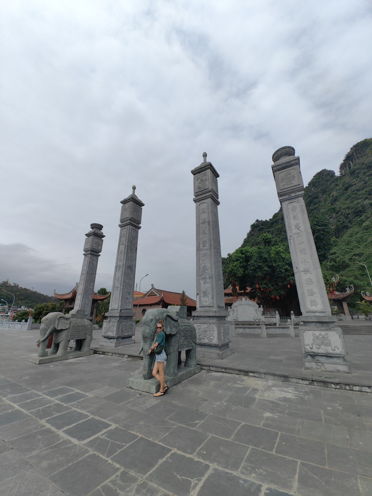 The image shows a woman standing next to a stone elephant statue, part of a monument in Hong Gai Ward, Vietnam. The monument consists of four tall stone pillars, each atop a stone elephant. The woman is wearing a green shirt, jean shorts, and sandals, and has sunglasses on. The sky is overcast. In the background, there are buildings with traditional Vietnamese architecture, as well as a green, mountainous landscape. The entire scene is built on a paved stone plaza.