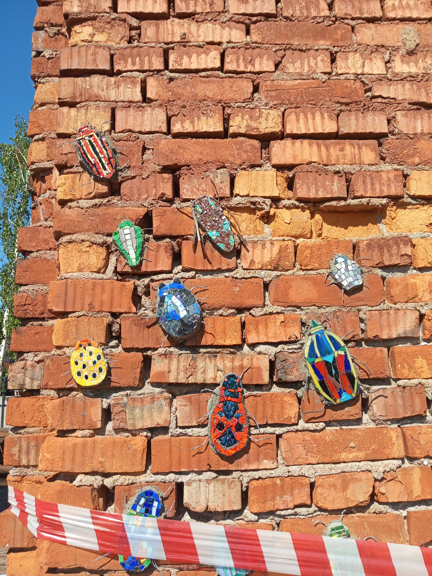 A close-up shot depicts a textured brick wall adorned with colorful mosaic insect art installations. The wall is made of weathered, red-orange bricks with uneven surfaces and mortar between them. Several mosaic-covered beetles are mounted on the brickwork at varying heights. These beetles are made of different colored tesserae and feature wire antennae and legs. A red and white striped caution tape runs diagonally across the lower section of the image, partially obscuring one of the mosaic insects. A tree branch with green leaves is visible on the left side of the frame. The sky appears blue in the top left corner. The image was taken in Saint Petersburg, Russia.