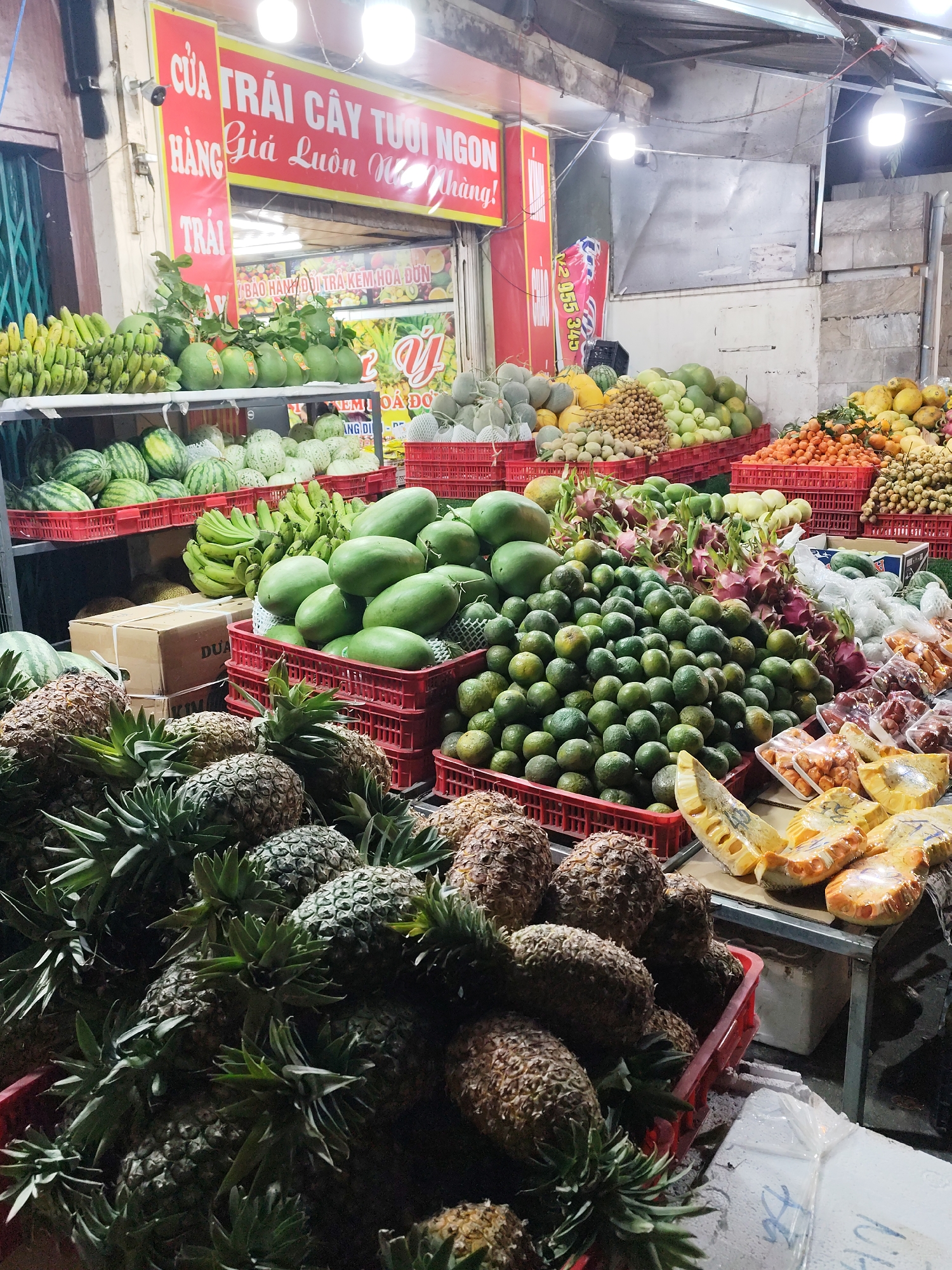 The image depicts a vibrant fruit market in Cam Ly - Đà Lạt, Vietnam. A large assortment of fruits are displayed in red plastic crates and on tables. Prominent fruits include pineapples in the foreground, along with watermelons, bananas, mangoes, citrus fruits, dragon fruit, longan, and packaged jackfruit. A sign reading "CỬA TRÁI CÂY TƯỜI NGON" (Fresh Fruit Shop) is visible.