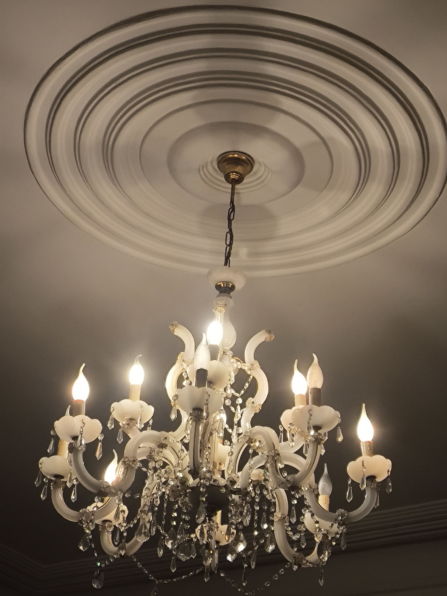 The image shows a close-up, low-angle shot of an ornate, multi-tiered crystal chandelier hanging from a ceiling adorned with a decorative plaster medallion. The chandelier is illuminated by faux candle-shaped bulbs and features numerous dangling crystal prisms and bead strings. The ceiling itself has a circular, stepped pattern created by the plasterwork. The lighting is warm, creating a soft glow and highlighting the reflective surfaces of the crystals. The background is a plain, dark ceiling. There are no people or animals visible in the image. The scene appears to be an interior, likely in a home or a venue like a restaurant or hotel. The location of Batumi, Georgia is not evident from the image itself.