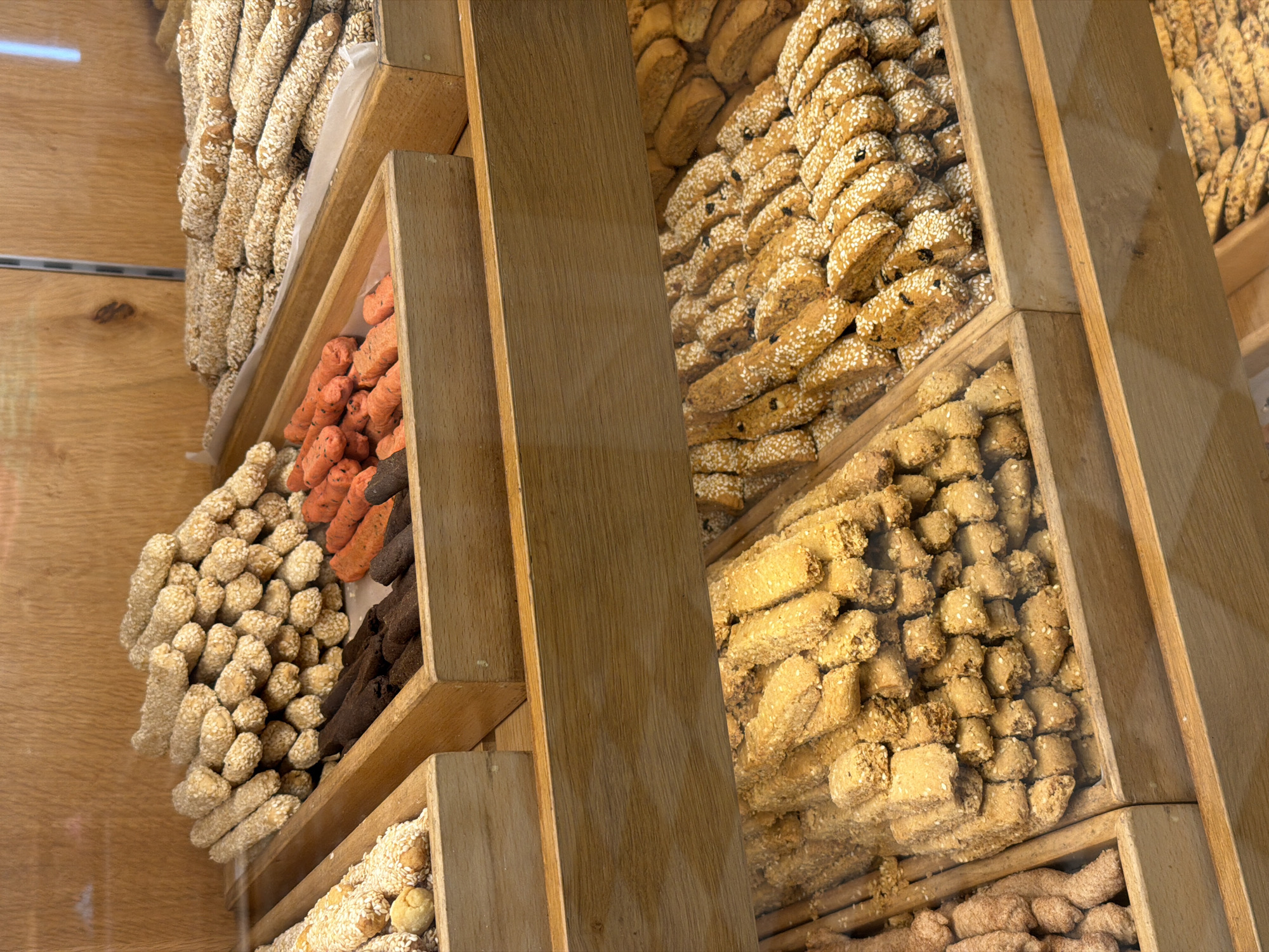The image shows a wooden display case filled with various types of cookies and biscuit sticks, seen behind a glass window. The display is arranged vertically in sections, each containing a different variety of baked goods. On the left side of the display, there are narrow, elongated biscuits covered in sesame seeds, alongside selections of orange and dark brown sticks. On the right, there is a variety of sesame-covered cookies with different shapes and sizes including bars and cubes. The wooden structure of the display case is prominent and appears to be made of light-colored wood. The image may be of baked goods in Hydra, Greece, as suggested by the prompt.