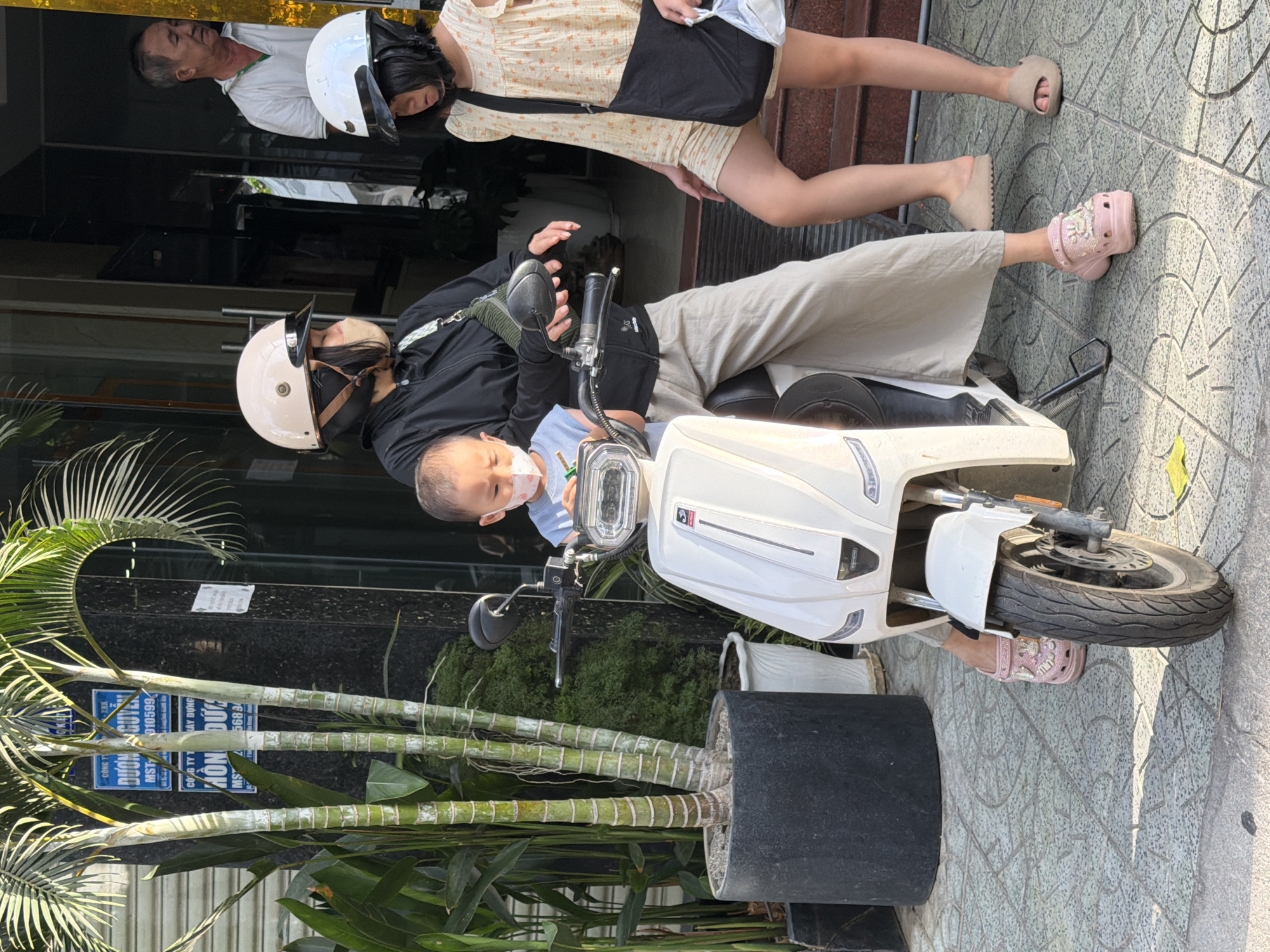 The image depicts a street scene in Nha Trang, Vietnam, with a focus on three individuals and a white electric scooter. The foreground features the front of the scooter, its wheel, and the tiled pavement. To the left, a large black planter holds several bamboo-like plants. In the midground, a woman wearing a white helmet and a light-colored face mask is seated on the scooter, holding a young child. The child, also wearing a light-colored face mask, is seated in front of her. The woman is dressed in a black jacket and loose-fitting beige pants. The child is wearing a light blue t-shirt. To the right of the scooter, a young girl wearing a white helmet and a light-colored dress stands with a black bag slung over her shoulder. She is wearing light pink sandals. Behind them, a man in a white polo shirt is partially visible in a doorway, looking towards the left. The background shows a building with a dark facade and signage in Vietnamese. The scene appears to be outdoors during daylight.