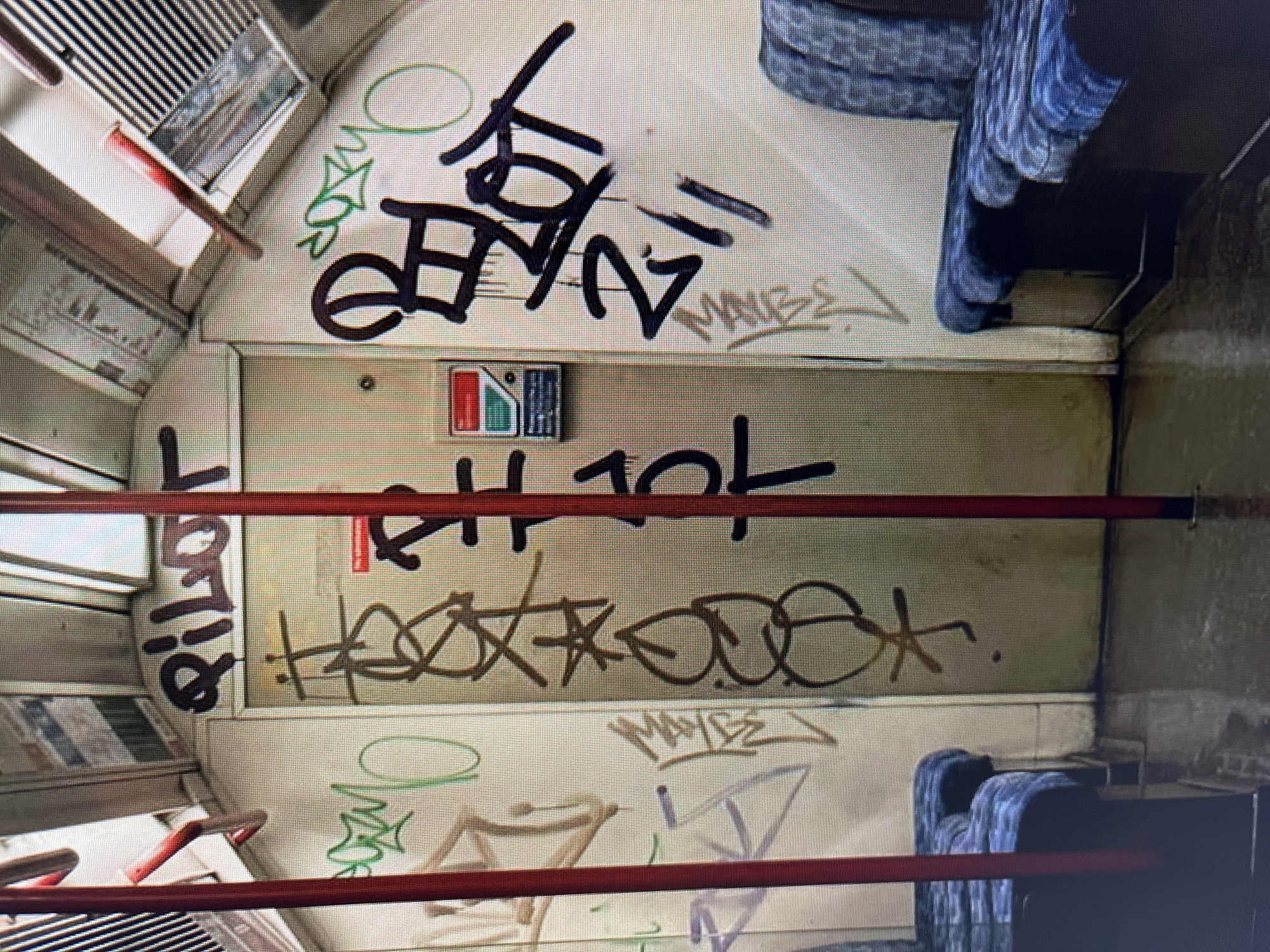The image shows the interior of a subway car, viewed from a high angle looking down. The car's interior is heavily adorned with graffiti, primarily in black and brown spray paint, with some green accents.
The subjects are the graffiti tags and the interior elements of the subway car. Objects include seats, handrails, ventilation grates, and a red and white indicator box on the wall. The scene is a typical, albeit vandalized, public transport environment. The setting is clearly a moving or stationary subway car.
No people are visible in the image, nor is any specific activity or event taking place beyond the presence of the graffiti.
Notable details include the extensive graffiti covering the walls and doors of the car. The graffiti features various tags and artistic styles. The lighting suggests an indoor environment, typical of a subway. The overall impression is one of neglect and vandalism.
Visible text includes various graffiti tags. On the left door, a dark tag appears to read "Pilot" with "Ollie" below it. Above the door, a larger black tag contains stylized lettering and what looks like "TBT". To the right of the door, there is a brown tag that is difficult to decipher but appears to have letters and symbols. Further down, a green tag is visible. A small red and white box on the door has some text or symbols on it, but they are too small to read.