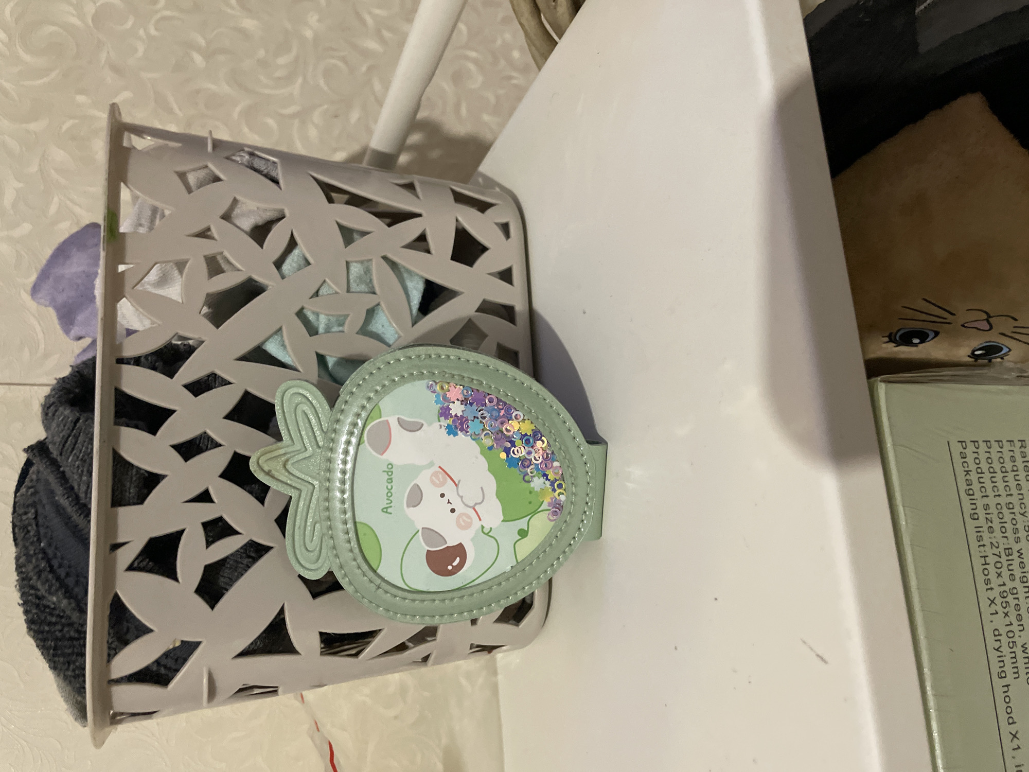The image presents a close-up view of a light gray plastic basket filled with clothing items. A green avocado-shaped object with cartoonish imagery rests against the side of the basket. Adjacent to the basket is a white surface, possibly a shelf or piece of furniture. A tan stuffed animal with cartoon eyes is also partially visible behind the white surface. The environment suggests an indoor setting, potentially a storage area or closet. The location in the city of Olginka, Russia is not evident from the details presented.