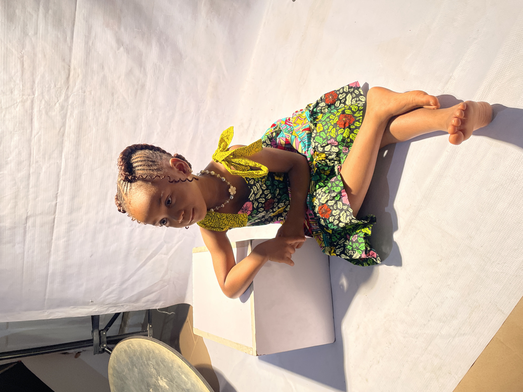 A young girl is lying on her stomach on a white backdrop. She is resting her forearms on a white box. The girl has braided hair and is wearing a colorful floral dress with a yellow sash. She is also wearing a beaded necklace. Her legs are bent at the knees. The girl is looking down at the box. A metallic object is in the lower left of the image. The backdrop appears to be set up indoors in Jos, Nigeria.