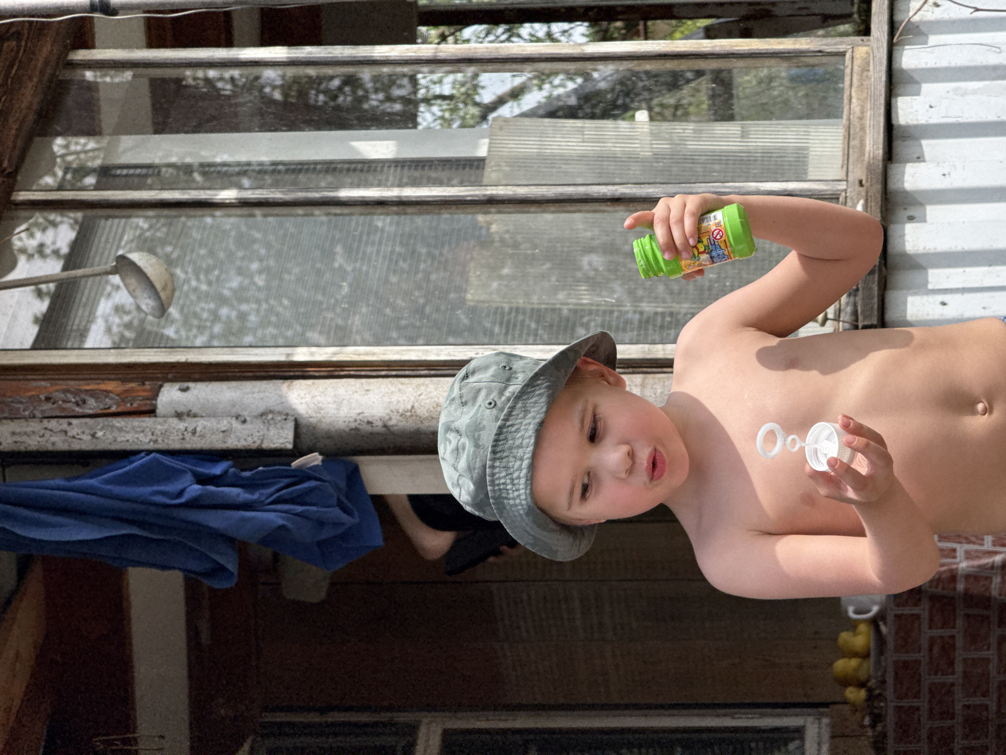 A shirtless child wearing a gray bucket hat blows bubbles outside a building. The child holds a green bubble solution container in their right hand and the bubble wand in their left, with two bubbles visible near their chest. The backdrop includes a window with a partial view of trees, a metal ladle, a blue cloth, and brickwork. The location is Chaikovsky, Russia.