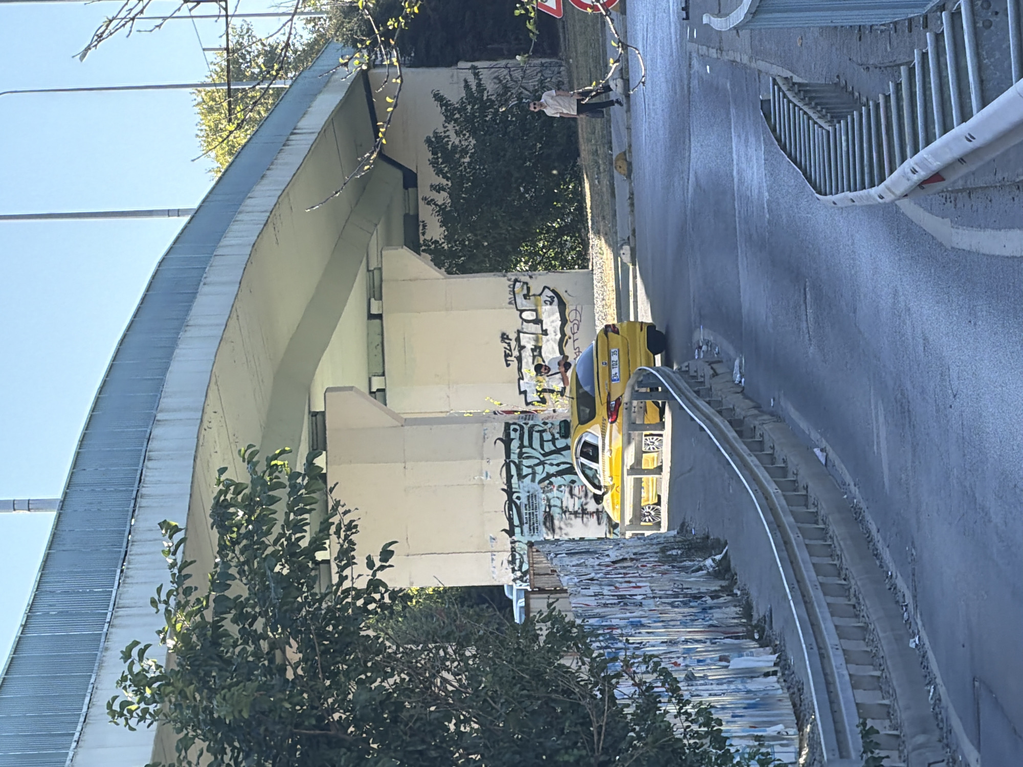 The image shows a street scene under an elevated road or bridge in Kadıköy, Turkey. A yellow taxi is parked or moving on the road, facing away from the camera. To the left, there is a metal guardrail and a wall covered with posters and papers. The bridge pillars are covered in graffiti. On the right side of the road, a man in a light-colored shirt and dark pants is walking away from the camera, carrying a bag. The sky is clear and blue, and there are trees and vegetation in the background. The overall impression is of an urban environment with elements of decay and urban art.