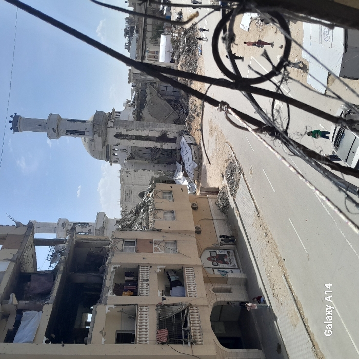 This image depicts a scene of extensive destruction in Khan Yunis, Palestinian Territory. The central focus is a significantly damaged mosque, identifiable by its distinct minaret and dome, both of which remain partially standing despite substantial collapse of the surrounding mosque structure. To the left, an adjacent multi-story building, appearing residential or commercial, is also severely damaged, with its upper floors partially collapsed, exposing rebar, debris, and interior spaces. Clothes are visible hanging on remaining balconies of this building.
A paved street runs across the mid-ground, flanked by sidewalks. On the left sidewalk, near the damaged building, a group of approximately three individuals stands near a shopfront displaying a "MARAH GAMES" sign. In the lower foreground, a dark-colored car is parked on the street, with a person in a green shirt standing nearby. To the right of the street, the area transforms into a expanse of dirt and rubble, where several more individuals, estimated at five to seven, are dispersed in small groups. Amidst this rubble, several white temporary tent-like structures are visible. The sky is partly cloudy blue, indicating daylight conditions. Metal railings and wires partially obstruct the immediate foreground view.
