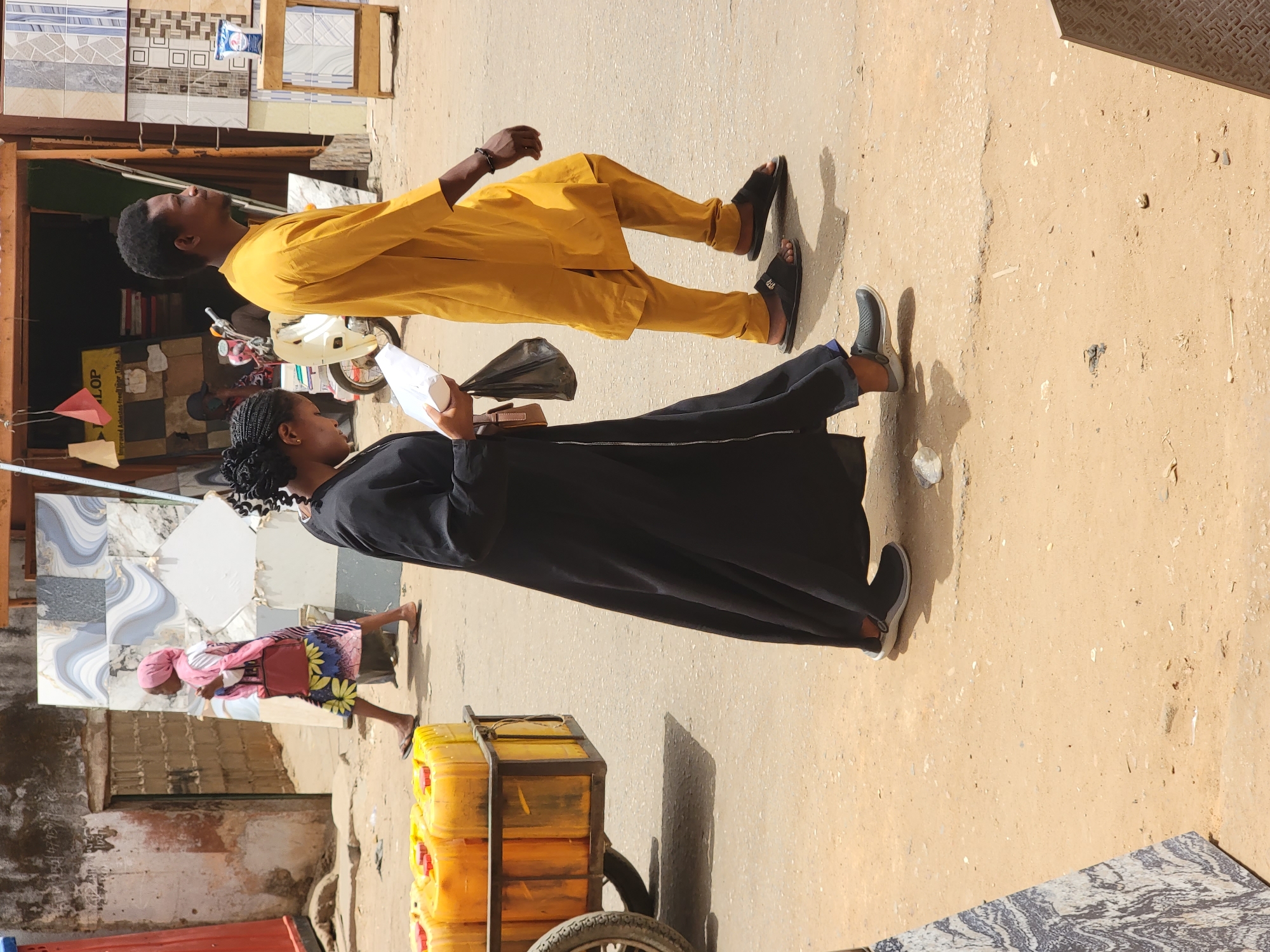 In a bustling street scene, a man dressed in a mustard-colored outfit strolls alongside a woman in a black abaya. The man wears dark sandals, while the woman sports gray sneakers. They are positioned on a sandy-colored pavement with a textured surface, potentially in Maiburiji, Nigeria. The woman holds a white paper and a dark bag in her hand. Adjacent to them, a metal cart laden with stacked yellow jerrycans rests on a pair of wheels. A woman wearing a pink head covering and colorful clothing walks into a building. Background elements include a building with tiles for sale and a parked motorcycle. The scene is illuminated by bright sunlight.