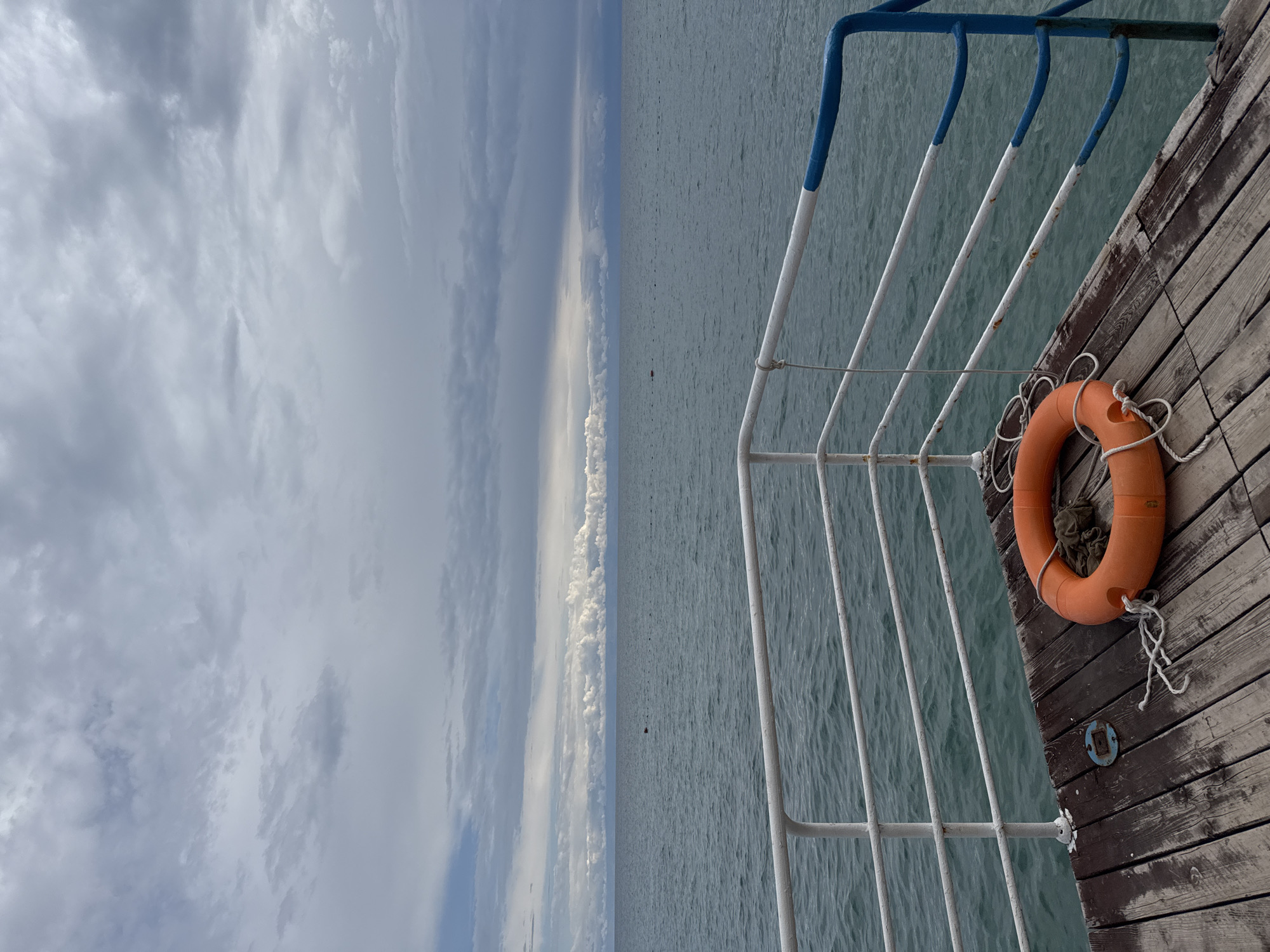 The image depicts a wooden pier extending over a body of water, likely Issyk-Kul Lake in Sary-Oy, Kyrgyzstan, under a cloudy sky. In the foreground, an orange life preserver is resting on the weathered wooden planks of the pier. A white railing, with sections painted blue, borders the pier. Beyond the railing, the tranquil, blue-green water stretches to the horizon, meeting a sky filled with layered grey and white clouds. The scene is serene and evokes a sense of a quiet day by the water. There are no people or animals visible in the image.