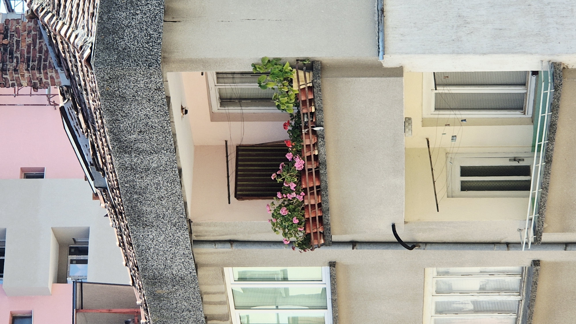 The image shows a close-up view of the facade of a multi-story building. In the foreground, there is a balcony with several potted plants, mostly with pink flowers, and one larger green plant. A gutter pipe runs vertically along the building's exterior. Above the balcony, the roof of the building is visible, made of terracotta tiles and partially damaged. A brick chimney is also present on the roof. Further in the background, other parts of the building with windows and balconies are visible, painted in pink and beige. The overall scene depicts an urban residential building in Lovech, Bulgaria, focusing on architectural details and plant life.