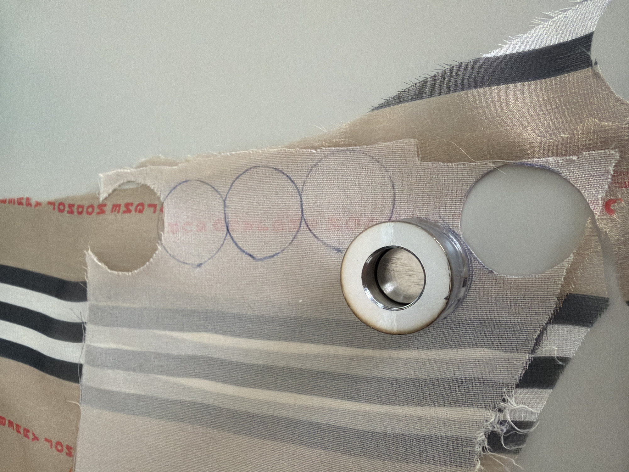 The media file captures a close-up view of textile work in progress, likely indoors on a light-colored surface, such as a table or workbench, in Saint Petersburg, Russia. The central subjects are several layered pieces of fabric being prepared for grommet installation.
One prominent fabric piece features horizontal stripes in varying shades of beige, light grey, and dark grey/black. Layered on top in sections is a lighter, possibly sheerer fabric, also with faint horizontal stripes. An underlying fabric, or the reverse side of one of the striped pieces, reveals red printed text, partially visible and inverted, which appears to read "LOEWE SOL" and "BERREY 70S."
The activity depicted is the marking and cutting of circular holes in fabric for grommets. Three blue-drawn circles are visible on the lighter fabric, indicating planned cuts, while one large circular hole is already cut out to the right. A silver-colored metal object, likely a part of a grommet setting tool or an actual grommet, is positioned near another cut-out circular opening. The edges of the cut fabric are frayed, characteristic of textile work. The lighting is even and diffused, suggesting a well-lit workspace. No people are visible.