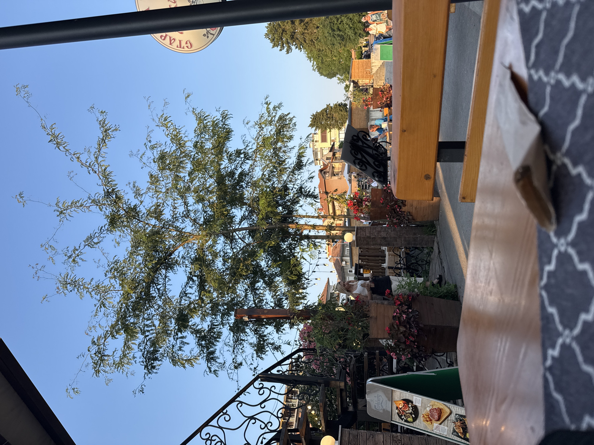 The image depicts an outdoor seating area of a restaurant or cafe, likely in the city of Primorsko, Bulgaria. In the foreground, a wooden table with a patterned tablecloth is visible, with a crumpled paper bag resting on it. A wooden bench is situated behind the table, partially obscuring a walkway. To the left, an ornate metal spiral staircase ascends, adorned with lights. Adjacent to the staircase is an A-frame sign displaying food items. Potted plants with red flowers and greenery are strategically placed around the seating area.
In the midground, several people are visible. A woman with blonde hair, wearing a white tank top and dark skirt, holds a glass of beer and stands near the staircase. Other individuals are seated at tables in the background. The environment appears to be a street or promenade, with buildings and trees lining the backdrop. A black lamppost stands on the right side of the image, partially obscuring a circular sign with red lettering. The sky is clear and blue, suggesting it is daytime, possibly late afternoon or early evening due to the warm lighting.