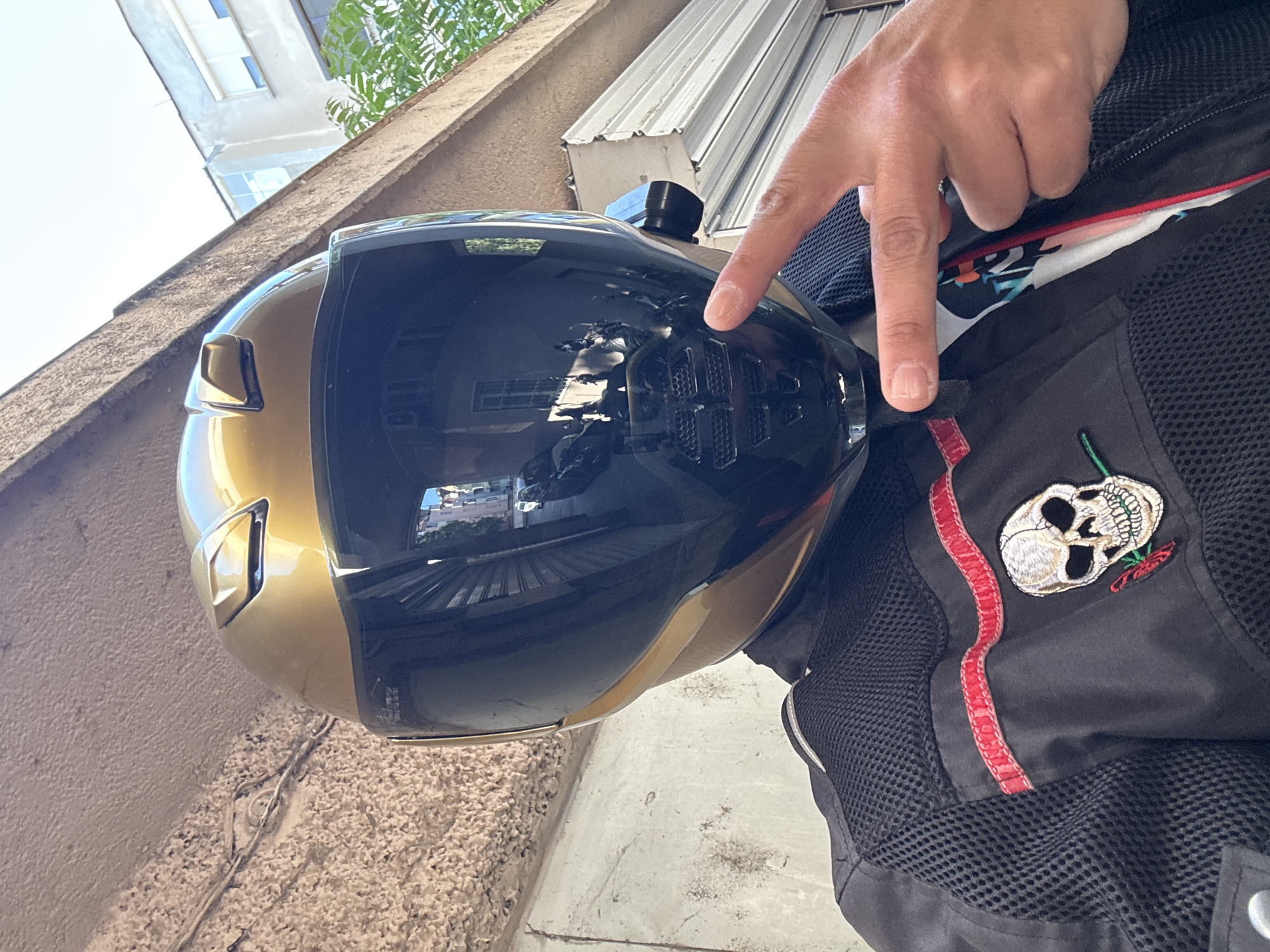 The image shows a close-up of a black and gold motorcycle helmet, with a person's hand pointing at the helmet's black visor. The helmet is reflecting the surroundings, including a building with windows and a roller shutter door. The person is wearing a black motorcycle jacket with a mesh panel and a skull and rose patch with a red stripe detail. The location is Plovdiv, Bulgaria, evident from the reflection in the visor, showing buildings typical of the city. The setting appears to be an outdoor urban area, possibly a street or parking area near buildings.