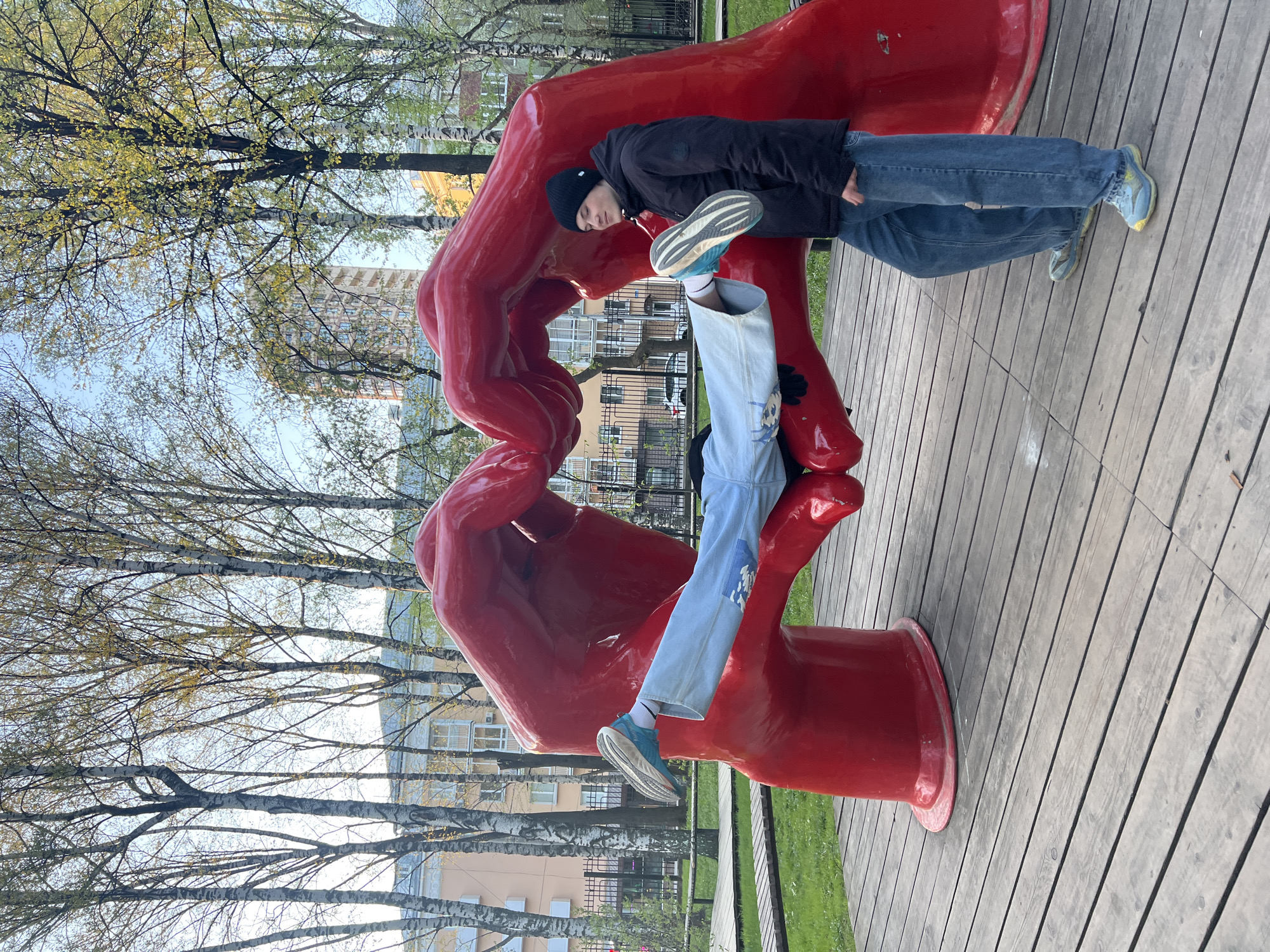 A person in a black jacket and blue jeans stands next to a large red sculpture of hands holding each other in a park setting. One leg extends through the sculpture, with a blue and white shoe visible. The park appears to be in an urban environment, with buildings and trees visible in the background. The scene is likely located in Perm, Russia, based on contextual information. The sculpture sits on a wooden platform.
