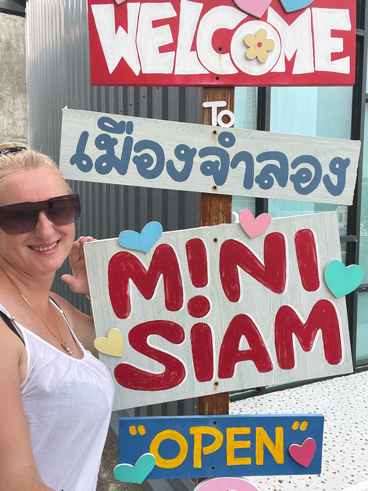 A woman stands in front of a series of signs. The topmost sign, a red rectangular board with white, hand-painted lettering, reads "WELCOME." Below this, a light blue sign with darker blue lettering, in Thai script, says "เมืองจำลอง" (Mueang Jamlong), which translates to "Mini Siam." Underneath this, a white sign with red, hand-painted letters displays "MINI SIAM," with a smaller blue sign below it that says "OPEN" in yellow. The signs are attached to a wooden post. The woman, with blonde hair and dark sunglasses, wears a white tank top and a gold necklace. Her left hand rests on the "MINI SIAM" sign, and she smiles at the camera. The background shows a corrugated metal wall and a tiled floor. The setting is consistent with the entrance area of Mini Siam, a popular tourist attraction in Pattaya City, Thailand, known for its miniature replicas of famous landmarks.