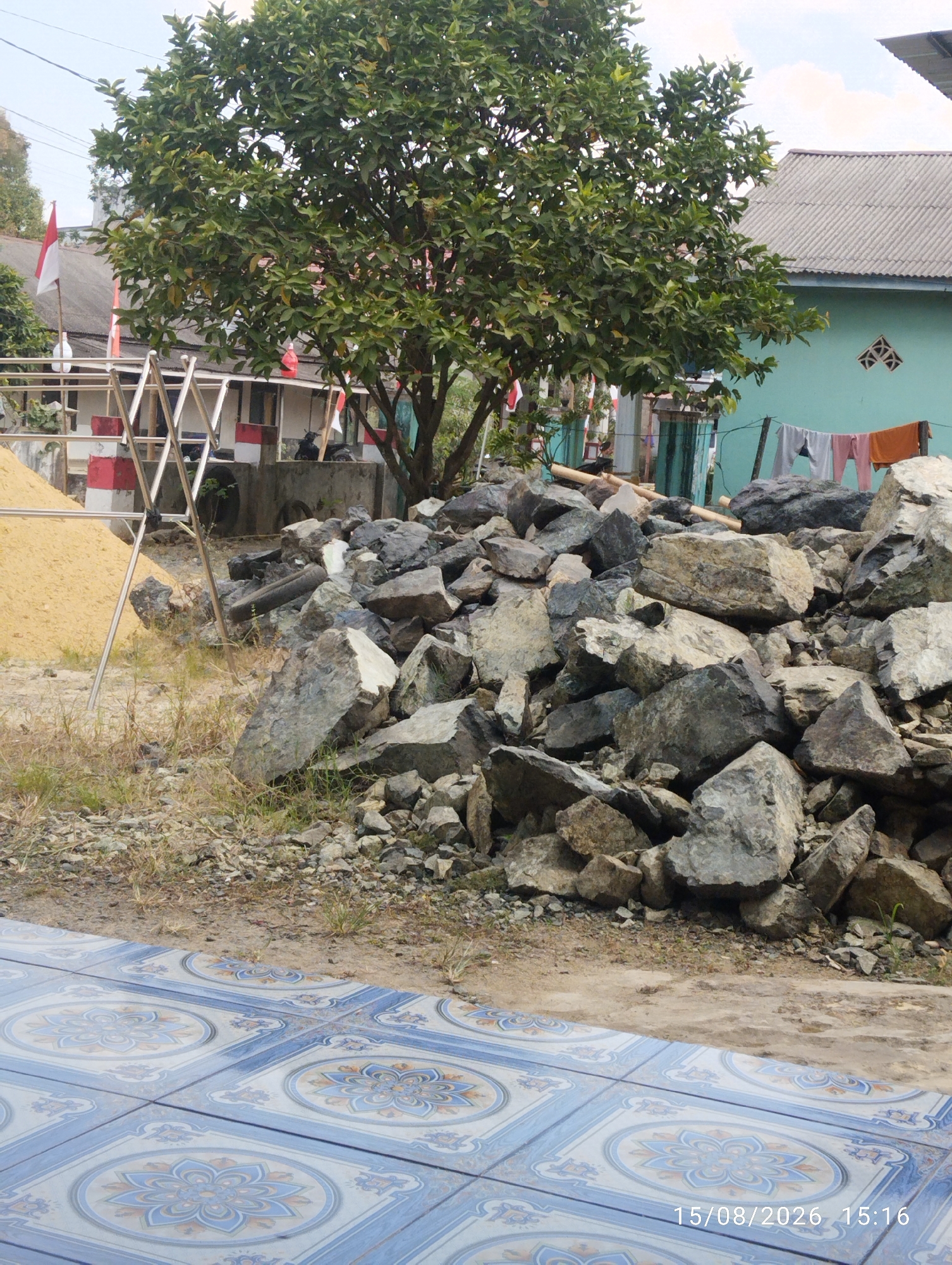 In the city of Tegal Rejo, Indonesia, an outdoor scene unfolds with a prominent tree and a pile of large rocks in the foreground. The tree, lush with green leaves, stands in the middle ground. Behind it, a turquoise building with a corrugated roof is visible, with laundry hanging on a clothesline. To the left of the tree, a white structure with red accents and metal framing is present, and a pile of yellow sand is also noticeable. The ground is a mix of dirt, gravel, and sparse vegetation. In the bottom portion of the image, a tiled floor with a blue and white floral pattern is visible. The date "15/08/2026 15:16" is superimposed on the lower right corner of the image.
