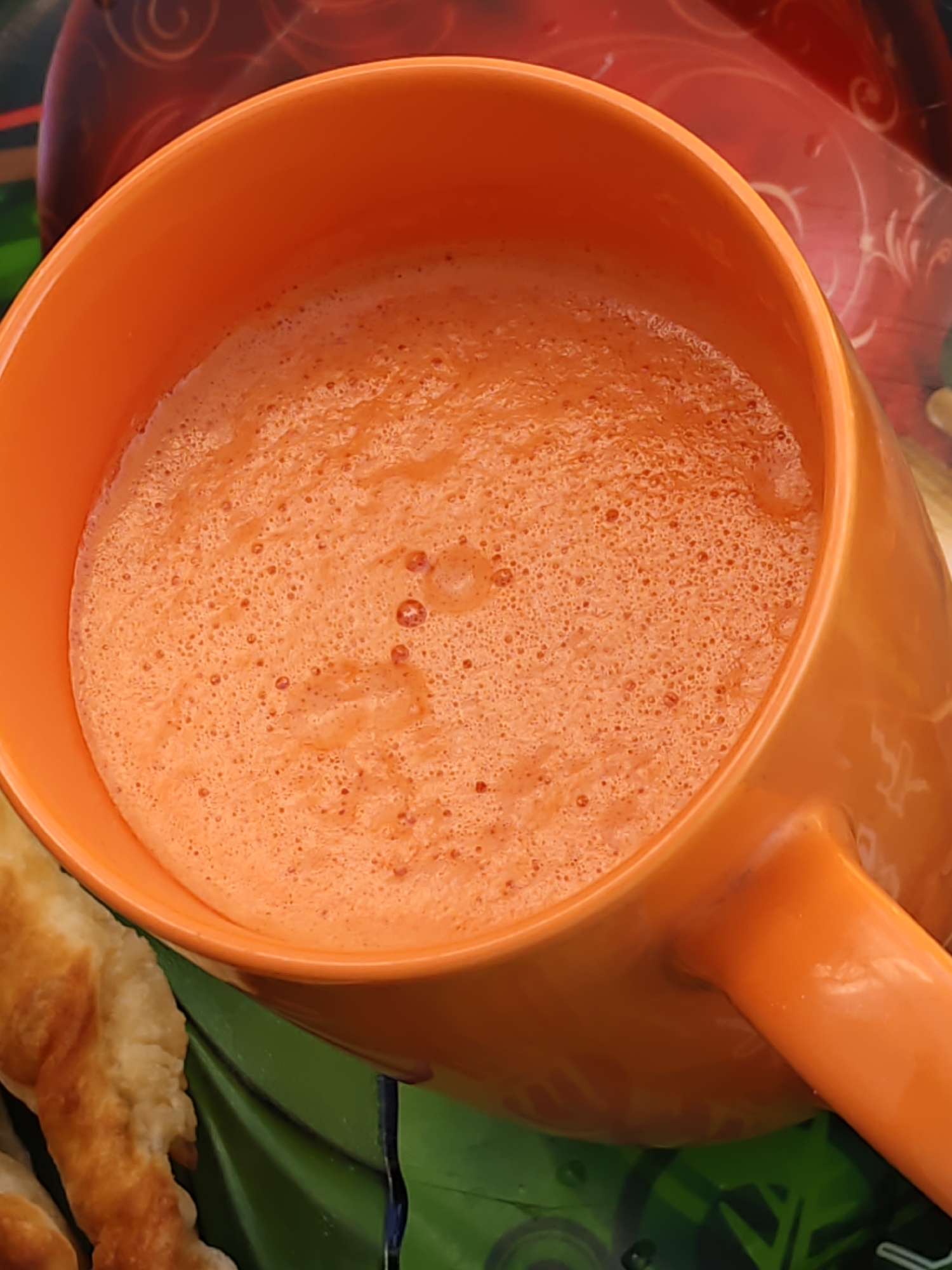 An orange mug filled with a frothy, orange beverage sits on a green surface with a patterned background. The mug has a handle. To the left of the mug are several golden-brown fried pastries. The lighting is bright, casting minimal shadows. The overall scene is a close-up shot of food and drink, with no discernible location or specific details related to Rostov-on-Don, Russia.