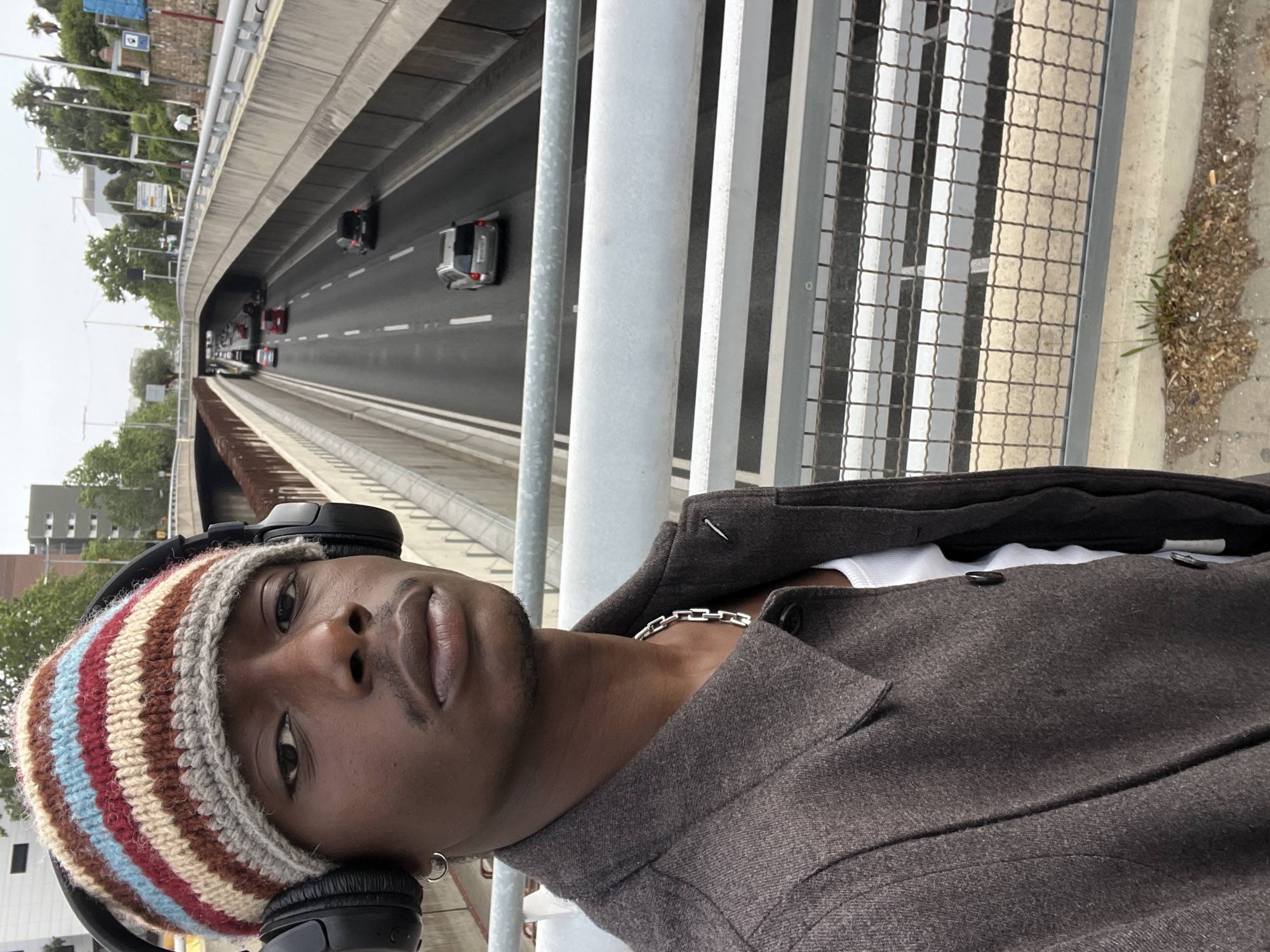 The original image is oriented 90 degrees counter-clockwise, depicting a person taking a selfie against an urban highway backdrop, likely in Barcelona, Spain. The main subject, a person with dark skin, is in the foreground, looking directly at the camera with a neutral to slightly serious expression. They are wearing a striped knitted beanie with colors including red, blue, cream, brown, and grey, along with black over-ear headphones. A dark grey or brown collared jacket is worn over a white undershirt, revealing a silver chain necklace and a small silver hoop earring.
The background features a multi-lane highway, possibly a sunken road or underpass, viewed from an elevated pedestrian bridge or overpass. Several cars, including sedans and potentially a truck, are visible on the road, indicating traffic. Metal railings and a mesh fence separate the person from the highway. Beyond the road, an urban landscape unfolds with numerous trees, including some palm trees, and distant buildings. The sky appears overcast, suggesting daytime with diffused light. On the ground near the railing, patches of weeds and scattered debris are visible. Streetlights and unreadable road signs are also present in the background.