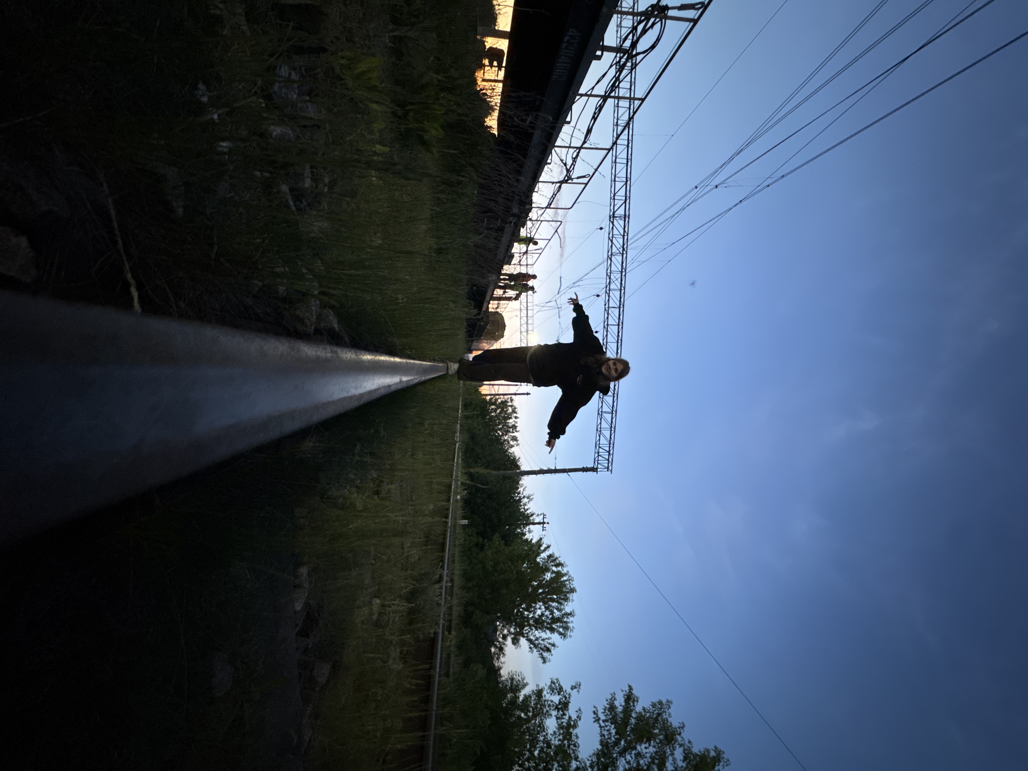 A wide-angle shot depicts a person standing with arms outstretched on a railroad track in Ферма, Russia. The perspective is skewed as if the camera is on the ground near the track. The person is wearing dark clothing. Overhead are power lines and a section of railroad infrastructure, potentially a bridge or elevated track. To the left, there is vegetation and the side of the railroad track. In the background, other people stand. The sky is visible, transitioning from a lighter blue near the horizon to a darker blue overhead. Trees are visible in the mid-ground.