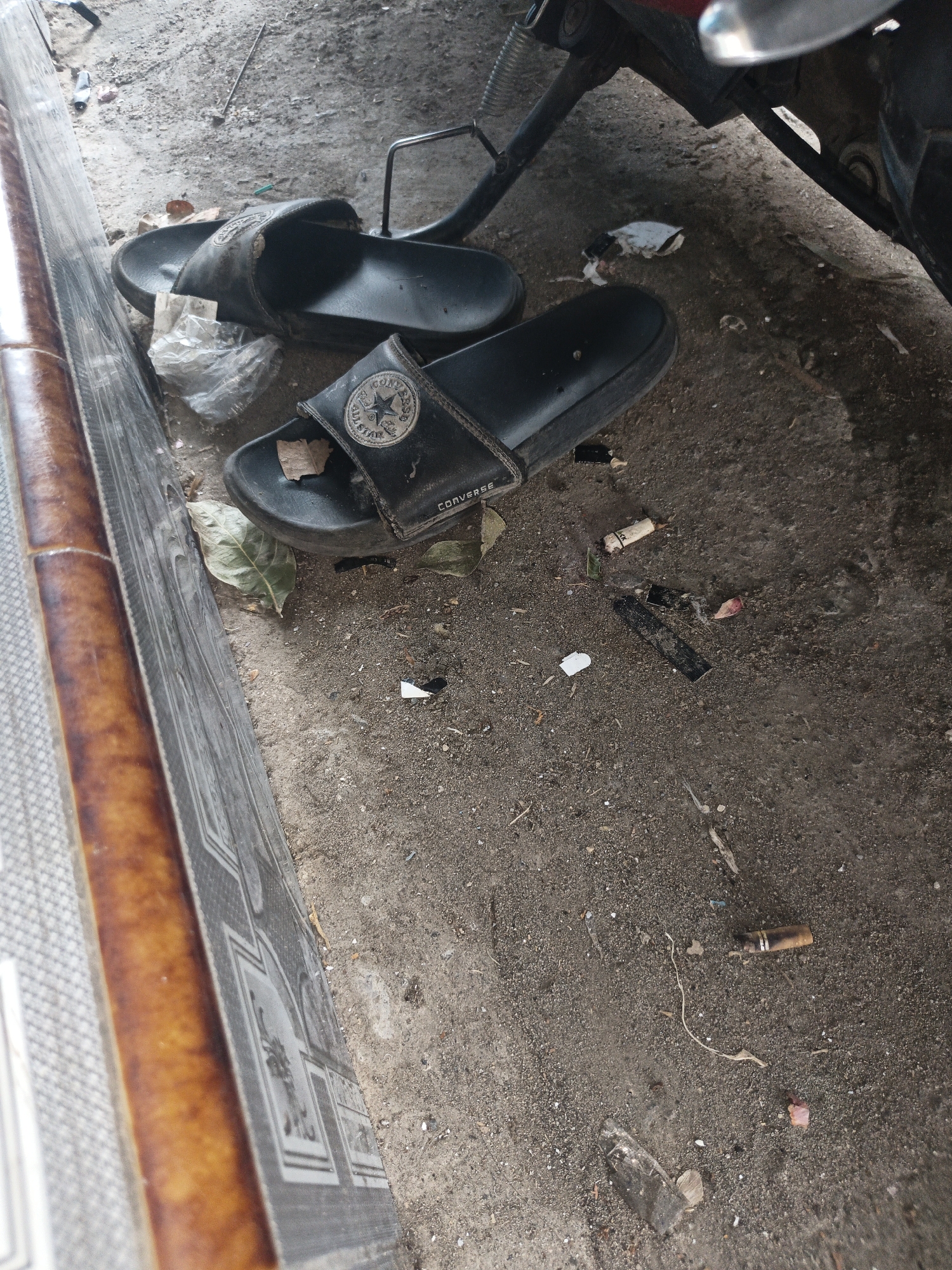 Two black Converse brand slide sandals are lying on a dusty, gritty surface. One sandal is partially obscured by a crumpled piece of plastic. The other sandal is more visible, with a circular Converse logo on the strap. The ground is littered with debris, including several cigarette butts, dried leaves, and small pieces of paper or plastic. The scene appears to be outdoors, possibly in a public area or alley, given the presence of discarded items and the general state of the ground. The angle suggests the photo was taken from a slightly elevated position, looking down at the sandals and the surrounding ground. The location is identified as Temuwulan, Indonesia.