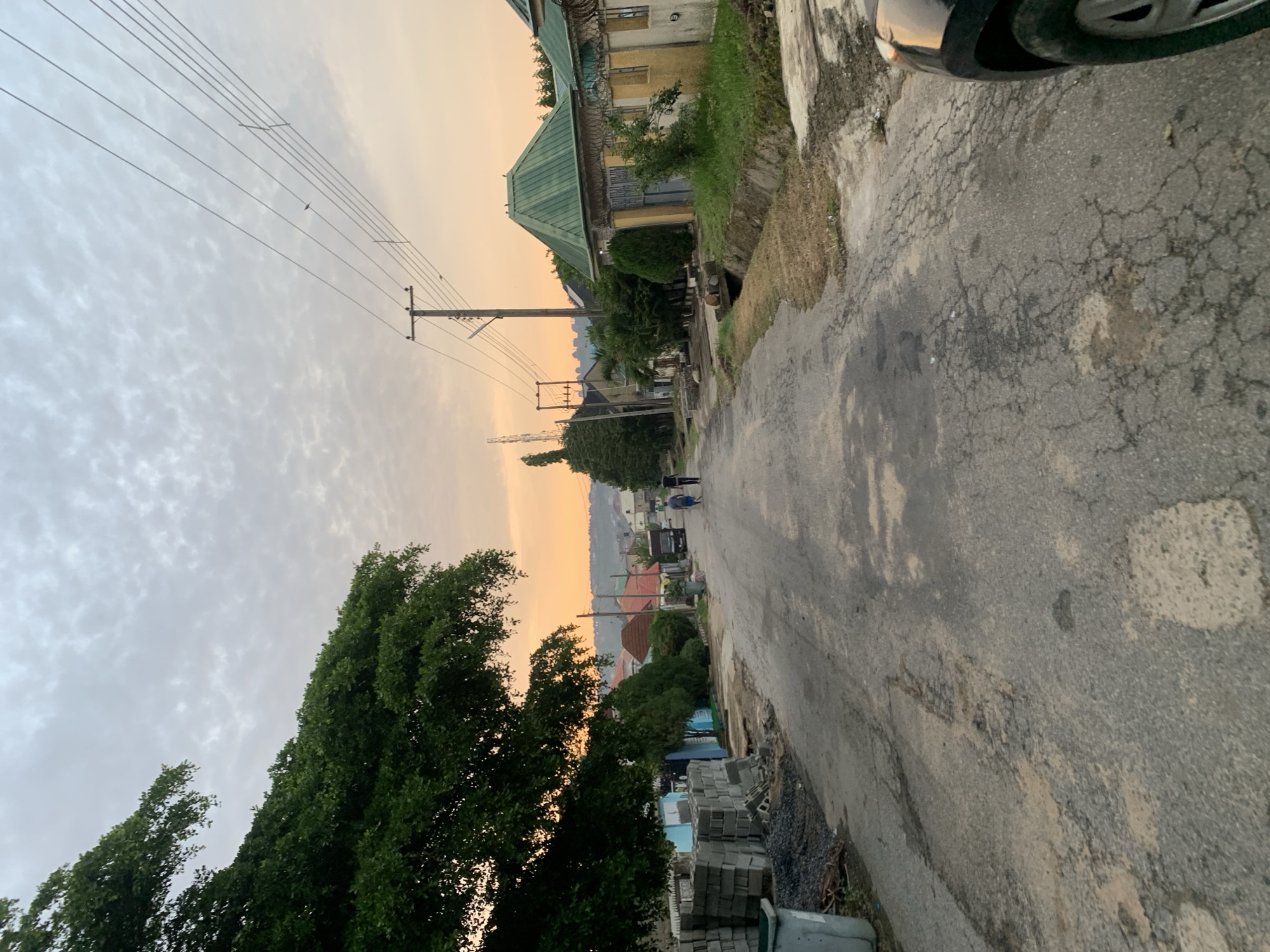The image depicts a street scene in Abuja, Nigeria, during either sunrise or sunset, as indicated by the warm, orange hues in the sky and the soft lighting. The road is paved but shows signs of wear, with cracks and patches. On the left side of the road, a large, leafy tree dominates the foreground, with its branches extending over the street. Adjacent to the tree, there are stacks of concrete blocks and a trash bin. Further down the street on the left, there are several houses and some smaller trees.
On the right side of the road, a row of houses with distinctive green roofs and yellow facades are visible. These houses appear to have high walls and gates, with some topped with barbed wire, suggesting a degree of security. There is also a drainage ditch running alongside the houses.
Two men are walking away from the camera down the center of the road, while in the distance, more houses and buildings are visible, stretching towards a slightly hazy horizon. Utility poles with overhead wires are present along the street, a common feature in urban and suburban environments. The overall impression is that of a residential area in a developing city, with a mix of construction materials, established homes, and ongoing infrastructure. The sky is partly cloudy, with patterns of clouds visible. The presence of a portion of a car in the lower right corner suggests the image was taken from within a vehicle or by someone standing near one.