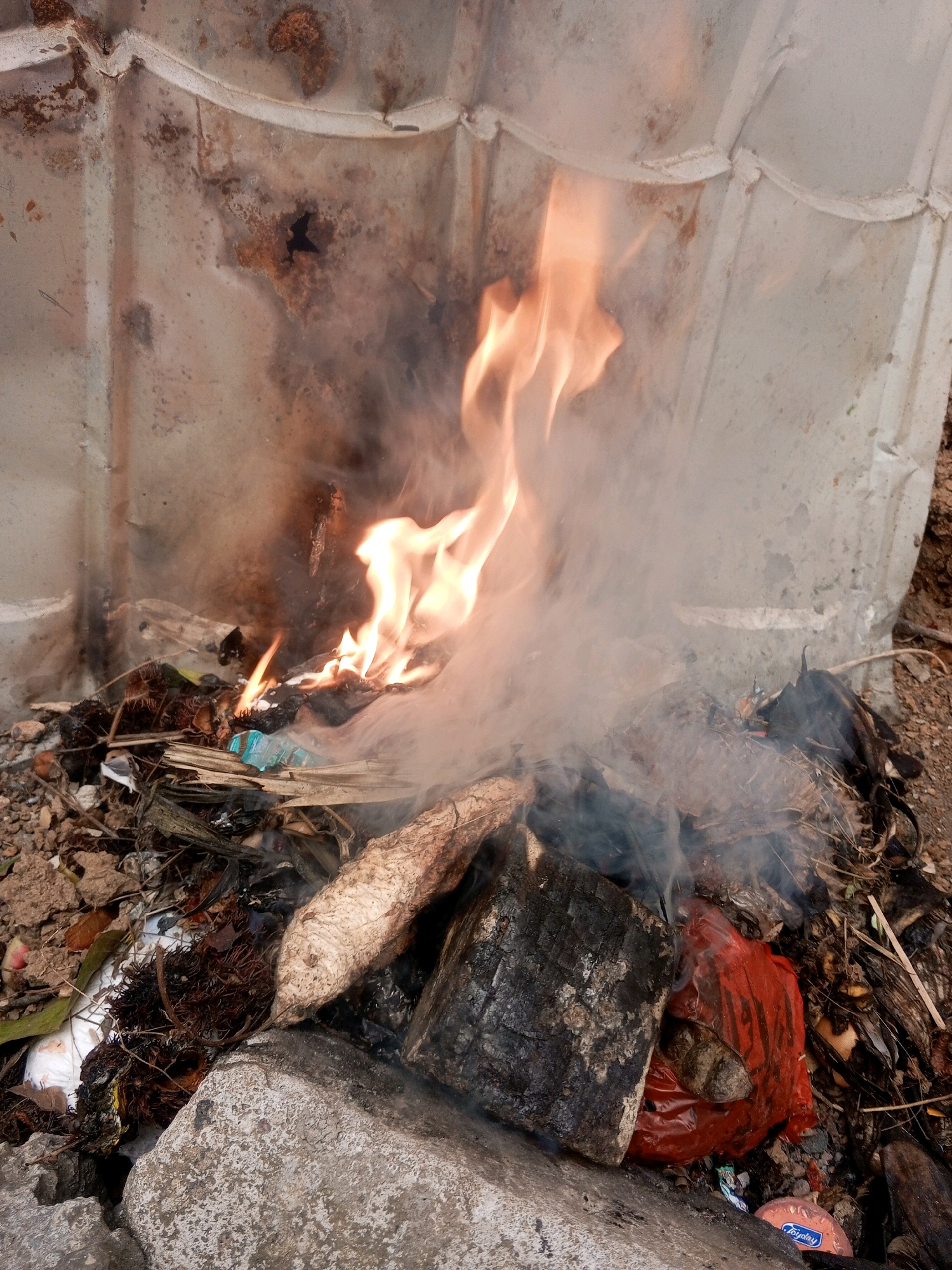 The image depicts an outdoor scene with garbage burning. The background consists of a corrugated metal structure, appearing rusted and weathered. In the foreground, a pile of miscellaneous debris, including dry leaves, twigs, and what appears to be a red plastic bag, is engulfed in flames. Smoke rises from the burning material. The ground is covered with soil, rocks, and more debris. The overall impression is one of waste disposal, likely in an informal setting. No people are visible in the image. The context of Cimarga, Indonesia, suggests a possible rural or semi-urban environment where such burning might occur.