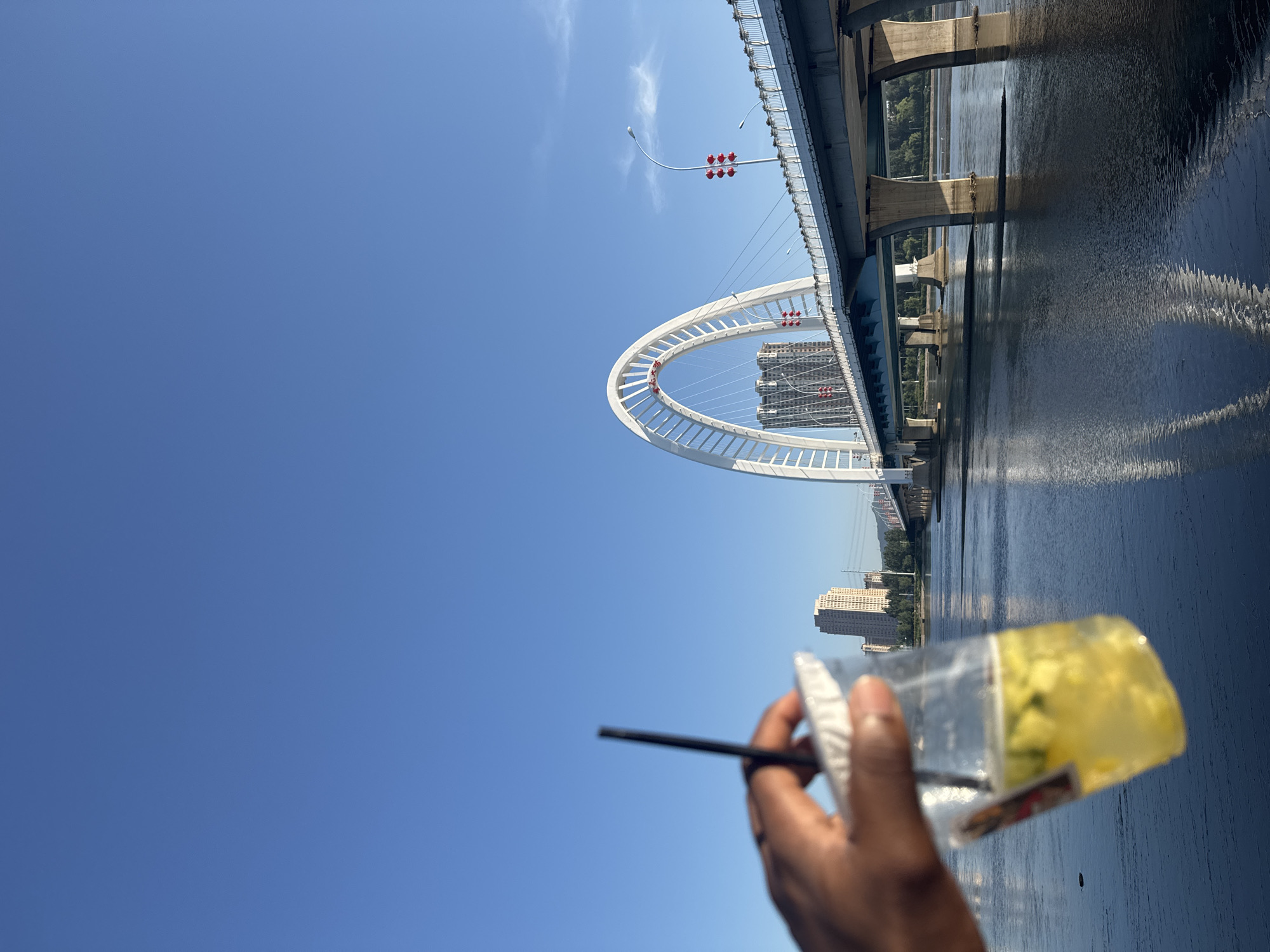 An overhead shot captures a hand holding a clear plastic cup filled with a yellow beverage and ice. The cup is positioned in the foreground, obscuring some of the background. Beyond the cup, a large modern bridge with an arching white structure dominates the scene. The bridge spans a wide body of water, its concrete pillars rising from the calm surface. In the distance, city buildings are visible, along with trees lining the banks of the water. The sky above is a clear, bright blue, with only a few wisps of clouds. The image appears to be taken on a sunny day, possibly in the city of Linghe, China, given the architectural style of the bridge.