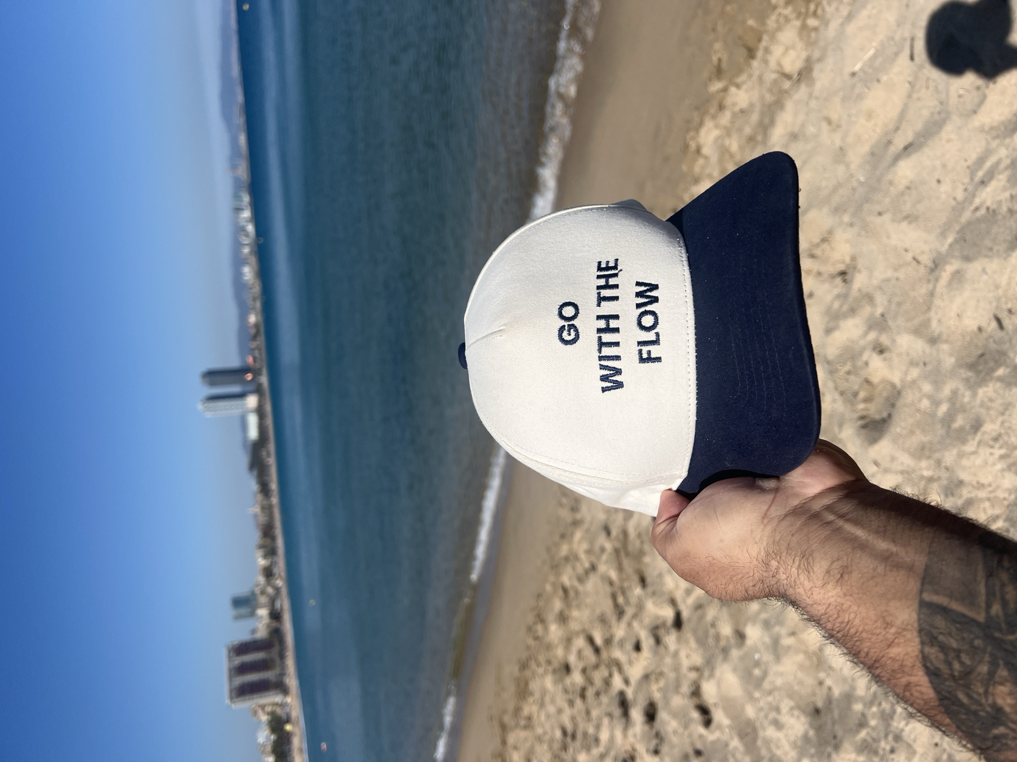A photograph shows a hand holding a white and dark blue baseball cap with the phrase "GO WITH THE FLOW" embroidered on the front. The background features a beach with sand, the ocean, and a cityscape in the distance. The photograph is taken during the day with clear blue skies and the water is a deep blue color. The sand appears light brown, and the beach looks clean and well-maintained. The cityscape is densely packed with buildings along the coastline, possibly resembling Barcelona's beachfront, though a definitive identification is difficult without more context.