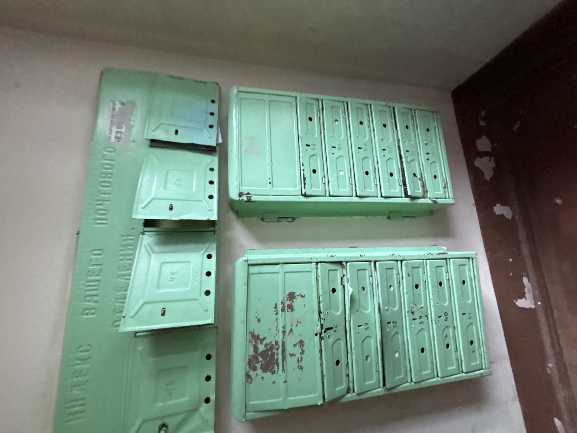 This image captures a wall in what appears to be an older residential building, featuring several mint green metal mailboxes. The mailboxes are arranged in a functional yet worn manner, showing signs of age and use.
On the left, a large, vertically oriented metal panel serves as a directory or information board. It is the same mint green color as the mailboxes and displays text in Cyrillic. The visible text on this panel reads "ИНДЕКС ВАШЕГО ПОЧТОВОГО ОТДЕЛЕНИЯ." which translates to "INDEX OF YOUR POSTAL DEPARTMENT."
To the right of this panel, there are two distinct units of individual mailboxes, stacked one above the other. Each unit contains multiple small, individual compartments, each with a small slot and a keyhole, characteristic of apartment mailboxes. The paint on these mailboxes is chipped in many places, revealing rust underneath, particularly on the bottom unit. Some mailbox doors are slightly ajar or appear to be damaged.
Numbers are visible on many of the individual mailbox doors, likely indicating apartment or box numbers. These numbers include 36, 37, 38, 39, 40, 41, 42, 43, 44, 45, 46, 48, and 50. The numbers appear hand-written or stenciled and are somewhat faded. The top unit has mailboxes numbered from approximately 42 to 50, while the bottom unit spans from about 36 to 41. The first compartment on the left of each horizontal unit appears larger or differently configured than the others.
The wall behind the mailboxes is light-colored, possibly off-white or cream, contrasting with a dark brown or maroon surface visible on the far right, which might be a door frame, another wall, or a corner. The overall impression is one of a utilitarian space in an older, perhaps Soviet-era, building. No people or specific activities are depicted.
