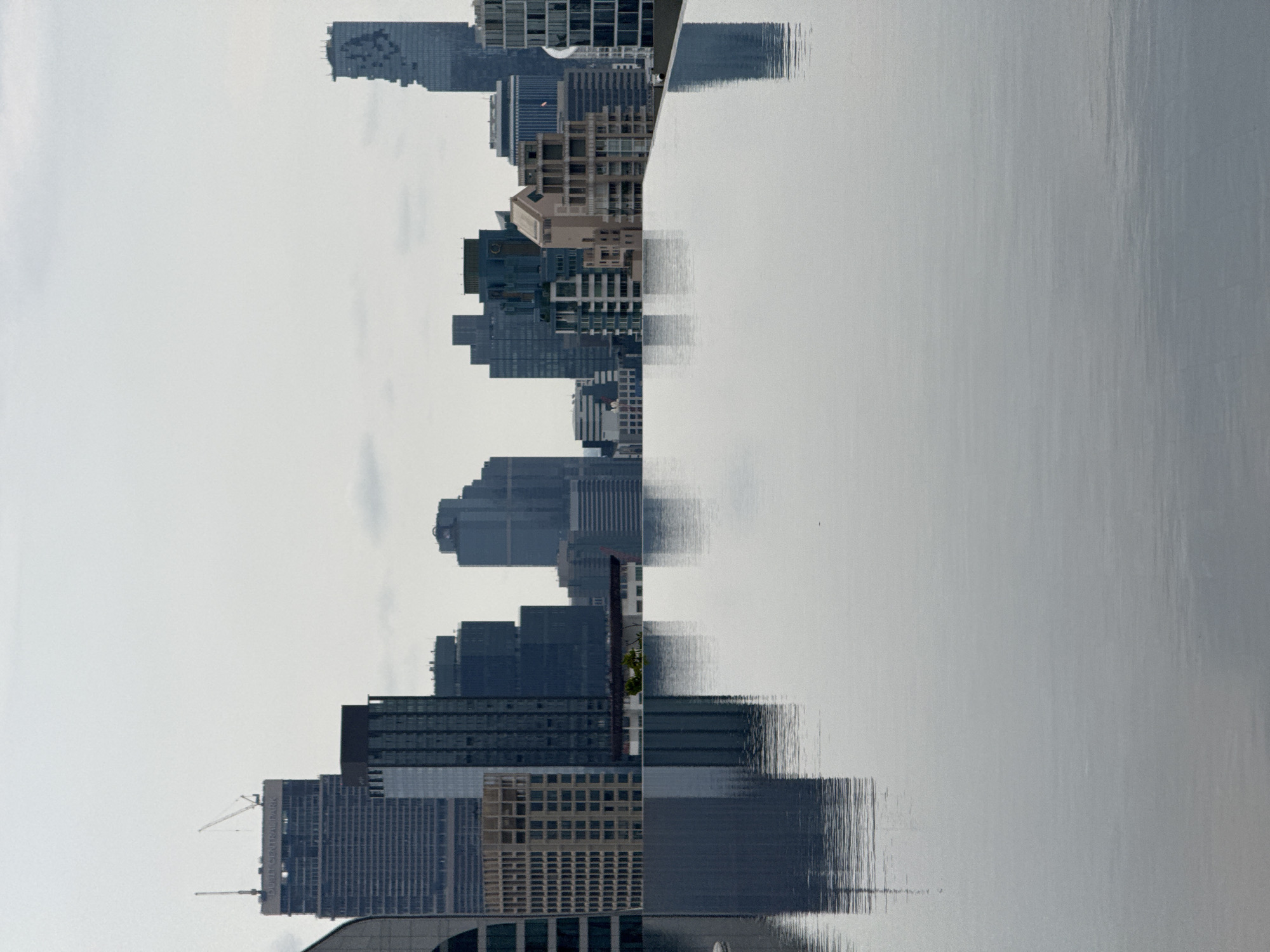 The image shows a reflection of Bangkok's skyline in water, likely the Chao Phraya River. The buildings are modern high-rises. The light is diffused, creating a hazy atmosphere. The reflection is slightly distorted by the water's surface. South Central Park is visible on a building. A crane is visible. The sky is overcast.