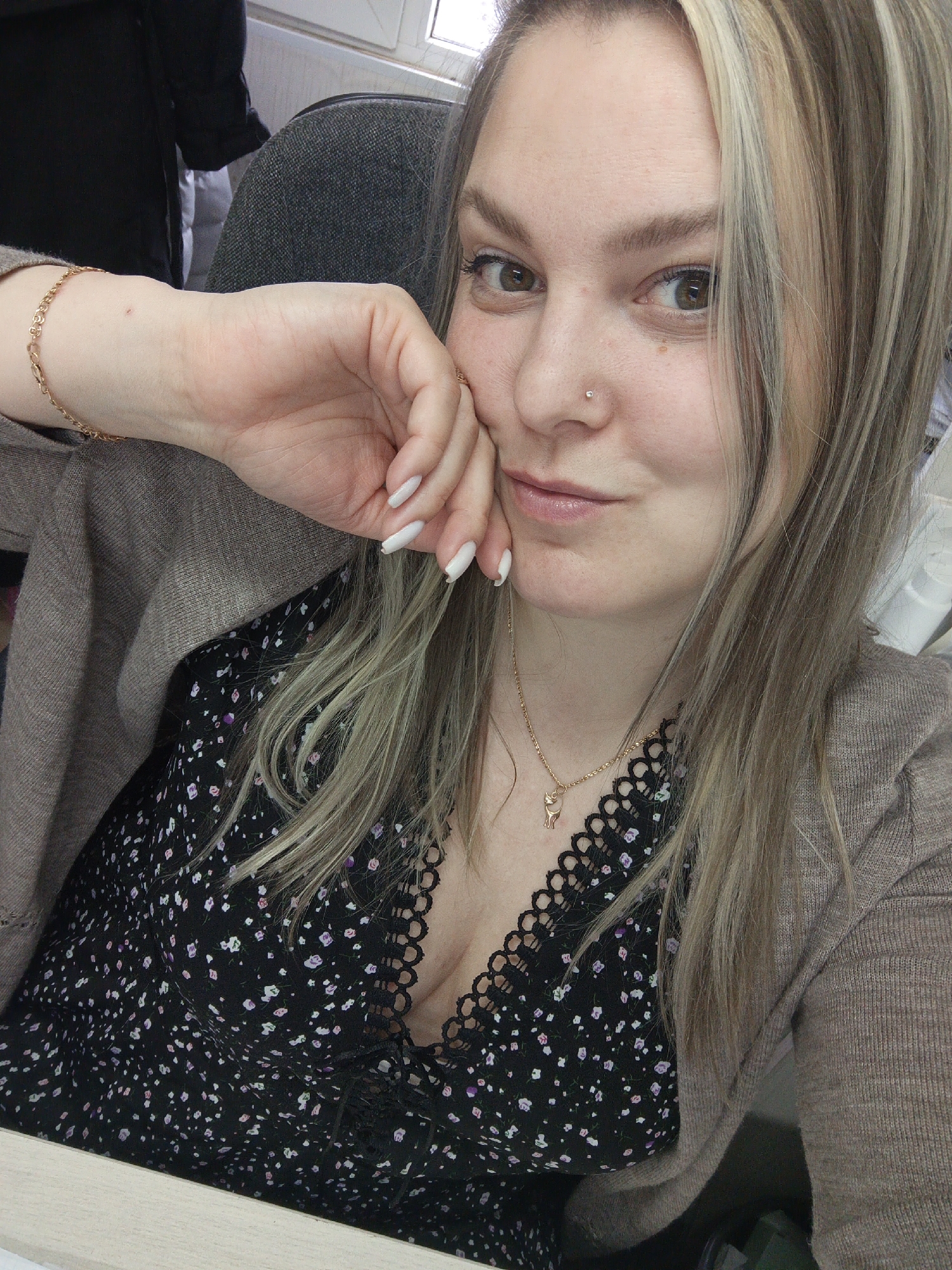 A woman is shown in a medium close-up selfie, appearing to be seated in an office setting. She has fair skin, light brown hair with blonde highlights, and brown eyes. A nose piercing is visible on her left nostril. She is wearing a black dress with a small floral pattern and lace detailing around the neckline, along with a tan cardigan. She also has a gold necklace with a small pendant and a matching bracelet. Her left hand is raised to her face, with long, white, manicured nails. The background includes a grey office chair and what seems to be a window. The setting is indoors with a light source coming from the window. The presence of Russian lettering on the side of a nearby piece of equipment indicates the city is Noviy, Russia.