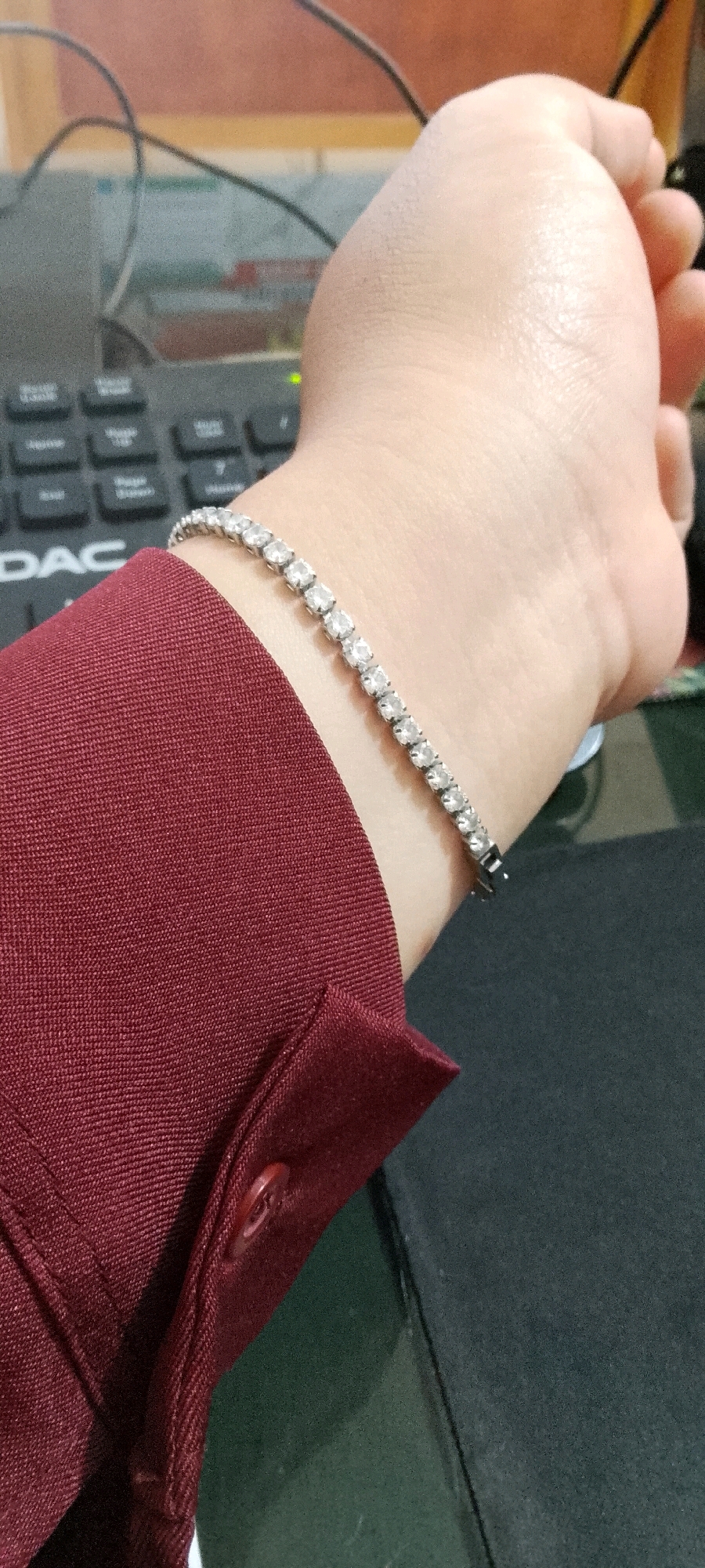 A close-up, indoor, medium-shot shows a person's left wrist and forearm, adorned with a tennis bracelet featuring small, clear, cubic zirconia stones set in a silver-toned metal. The wrist is bent at a slight angle, and the hand is partially visible, curled inwards. The person is wearing a dark maroon button-up shirt, with the collar and a visible button highlighted. In the background, a portion of a black keyboard is visible, with some keys obscured. The environment appears to be a workspace or office, with a hint of a dark green surface to the right of the frame, potentially a desk or another object. The lighting suggests an indoor setting, possibly under artificial light. No specific location in the city of Unknown, Indonesia, can be identified from the visual information provided.