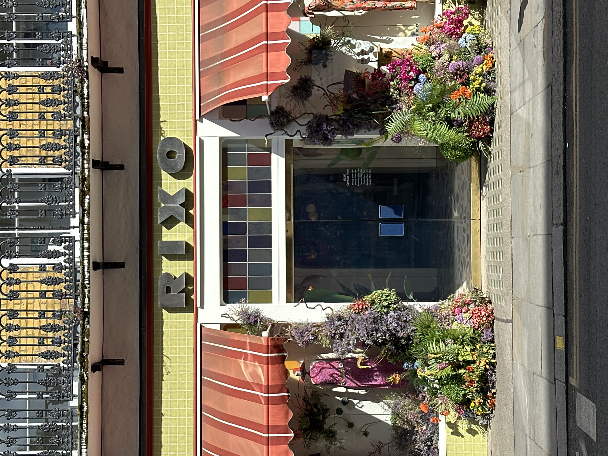 The image depicts the exterior of a store named "RIXO" located in London, United Kingdom. The store front is adorned with colorful flowers and plants on either side of the entrance. The store's sign is vertically aligned, and awnings with red and white stripes are above the windows. The door is a glass double-door. The building's facade features yellow tiles and a decorative iron balcony on the upper levels. The sidewalk and street are visible in the foreground, indicating an urban setting.