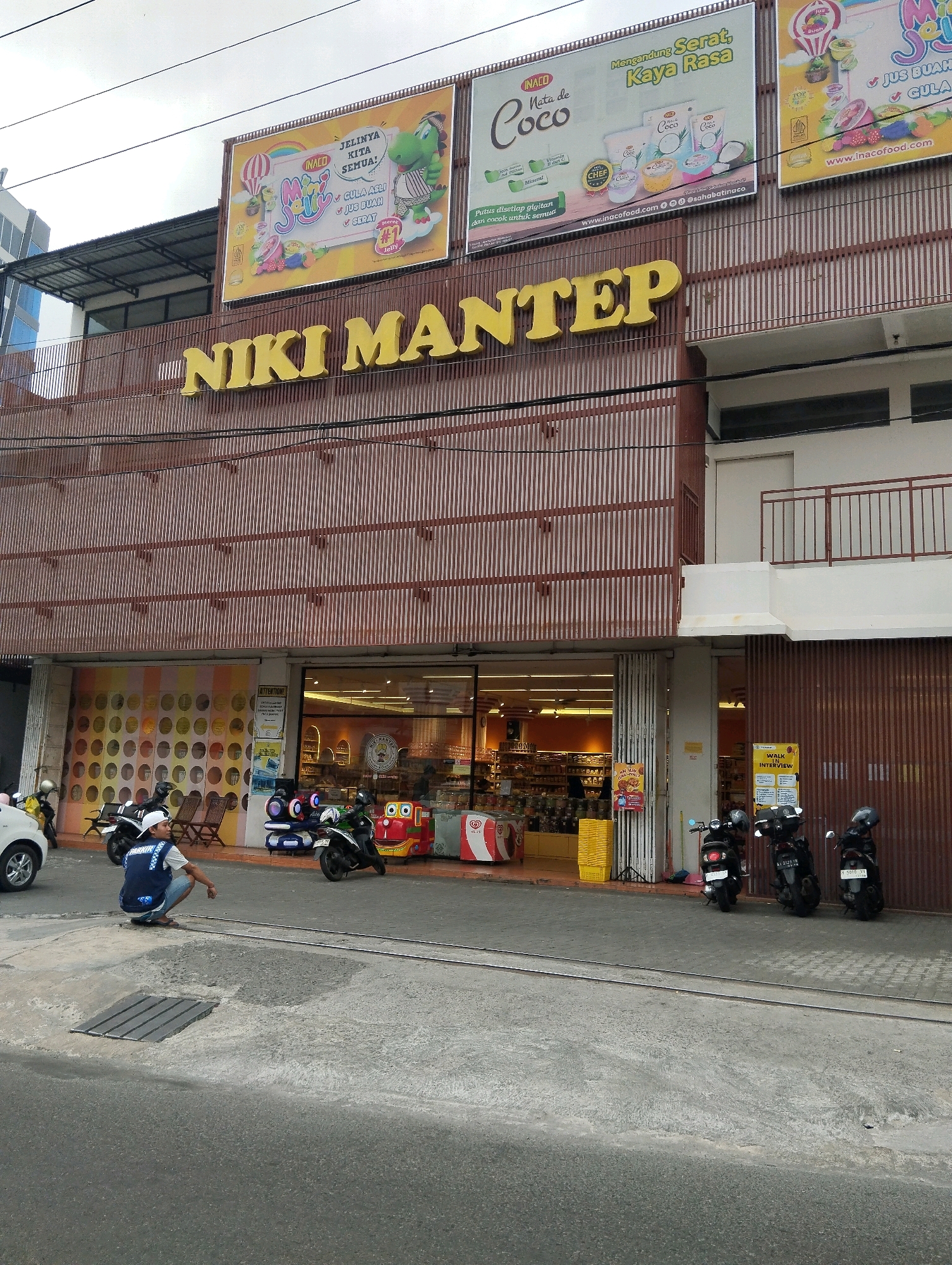 The image shows a storefront in Salatiga, Indonesia. The building has a prominent sign that reads "NIKI MANTEP" in large yellow letters. Above the sign, there are three billboards advertising various products, including what appears to be snacks and drinks. The storefront itself features large glass windows, revealing shelves stocked with merchandise inside. Several motorcycles are parked in front of the store, and a person in a blue vest is crouching on the sidewalk. The street in front of the building is paved, and there are some metal rails embedded in the ground. The sky is overcast.