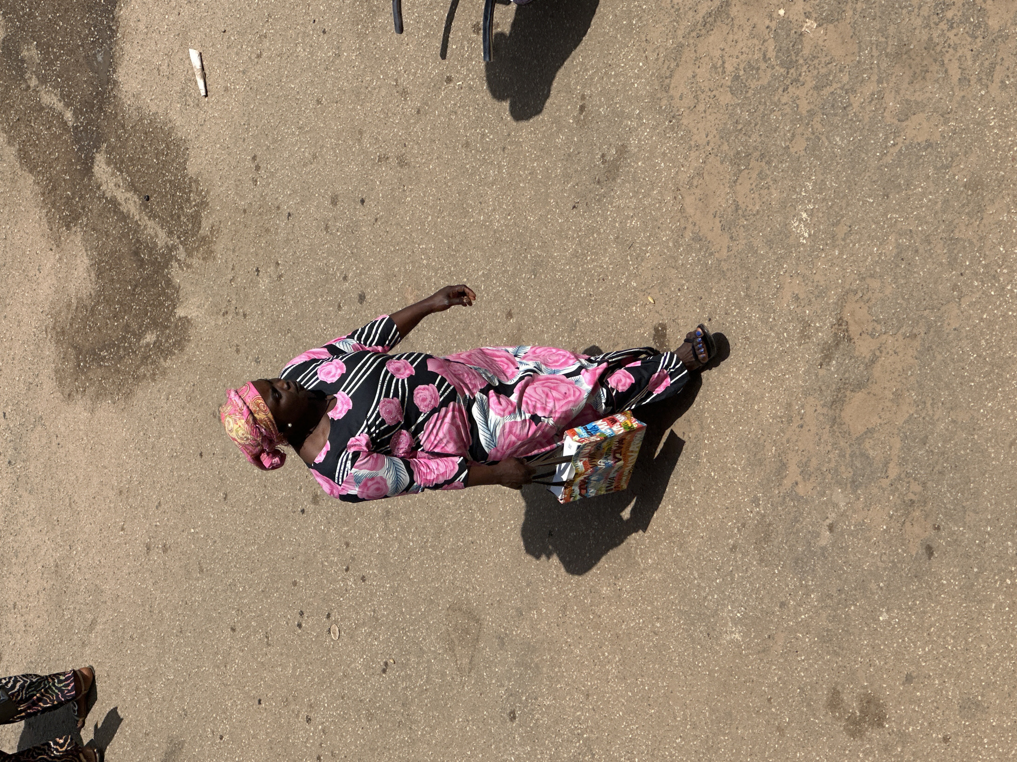 The overhead shot captures a woman walking on a concrete surface, likely in Bukuru, Nigeria. She is dressed in a vibrant, floral-patterned black and pink dress and a matching pink headscarf. The dress features large pink roses with black and white striped accents. Her right hand holds a colorful, rectangular object, possibly a bag or a collection of items, with visible text in various colors. She is wearing dark sandals.
The ground is a textured, light brown concrete with some damp patches and scattered debris, including a small white object resembling a rolled piece of paper near the top left. Shadows suggest the sun is casting strong light, indicating it's likely daytime, perhaps late morning or early afternoon. The overall scene is mundane, depicting everyday movement on a street or walkway. There are no overt emotional cues visible due to the overhead perspective and focus on the individual. Other figures are partially visible at the bottom of the frame, also walking.