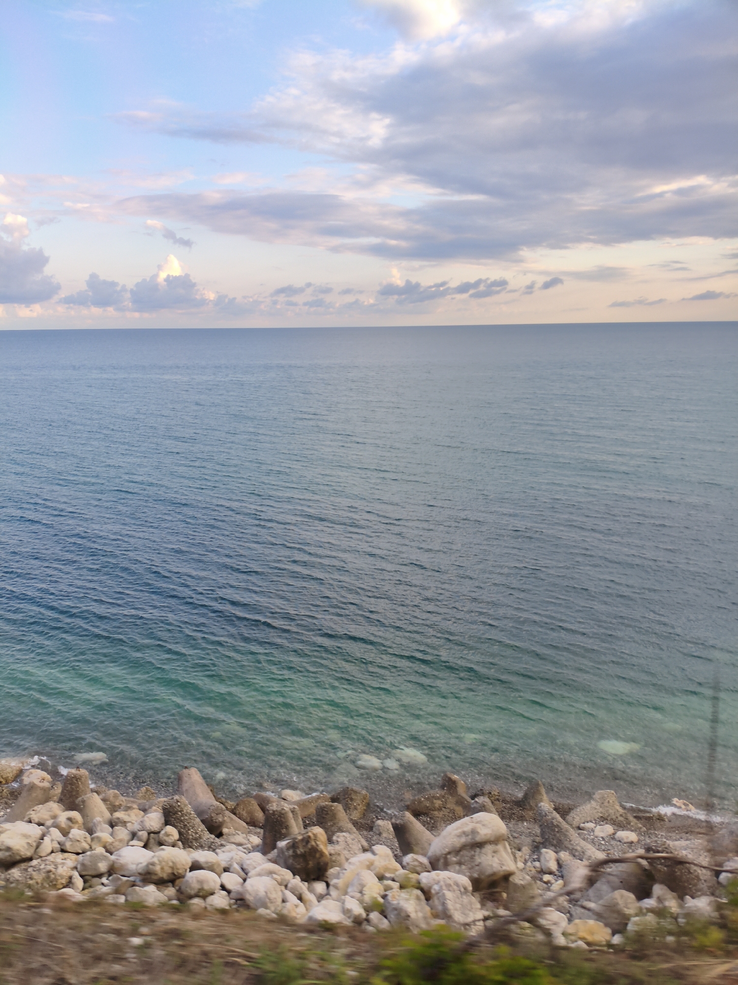 The image depicts a coastal scene in Chigripshi, Abkhazia. The horizon separates a vast expanse of the Black Sea from a cloudy sky. The sea is a deep blue, transitioning to a lighter, turquoise hue near the shore. Along the coastline, a collection of large, irregularly shaped rocks and concrete tetrapods are visible, serving as a breakwater. The foreground shows a narrow strip of earth with sparse vegetation. The overall impression is one of a natural, somewhat rugged coastline.