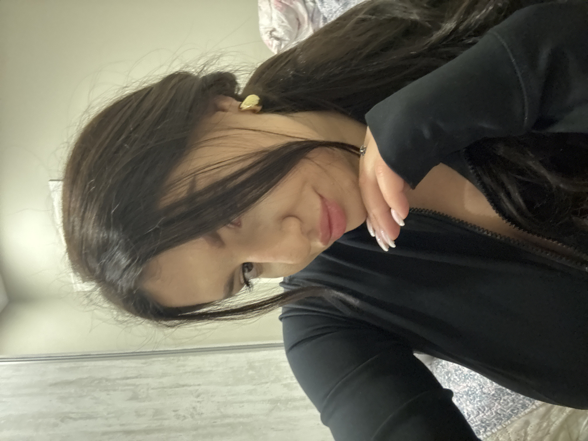 A woman with long, dark hair partially obscures her face, looking down. She wears a black, zip-up garment. Her fingernails are long and white. A gold earring is visible. The background includes a light-colored wall, a patterned bedspread, and a sliding door. There is no evidence to suggest the location of the image is Burgas, Bulgaria.